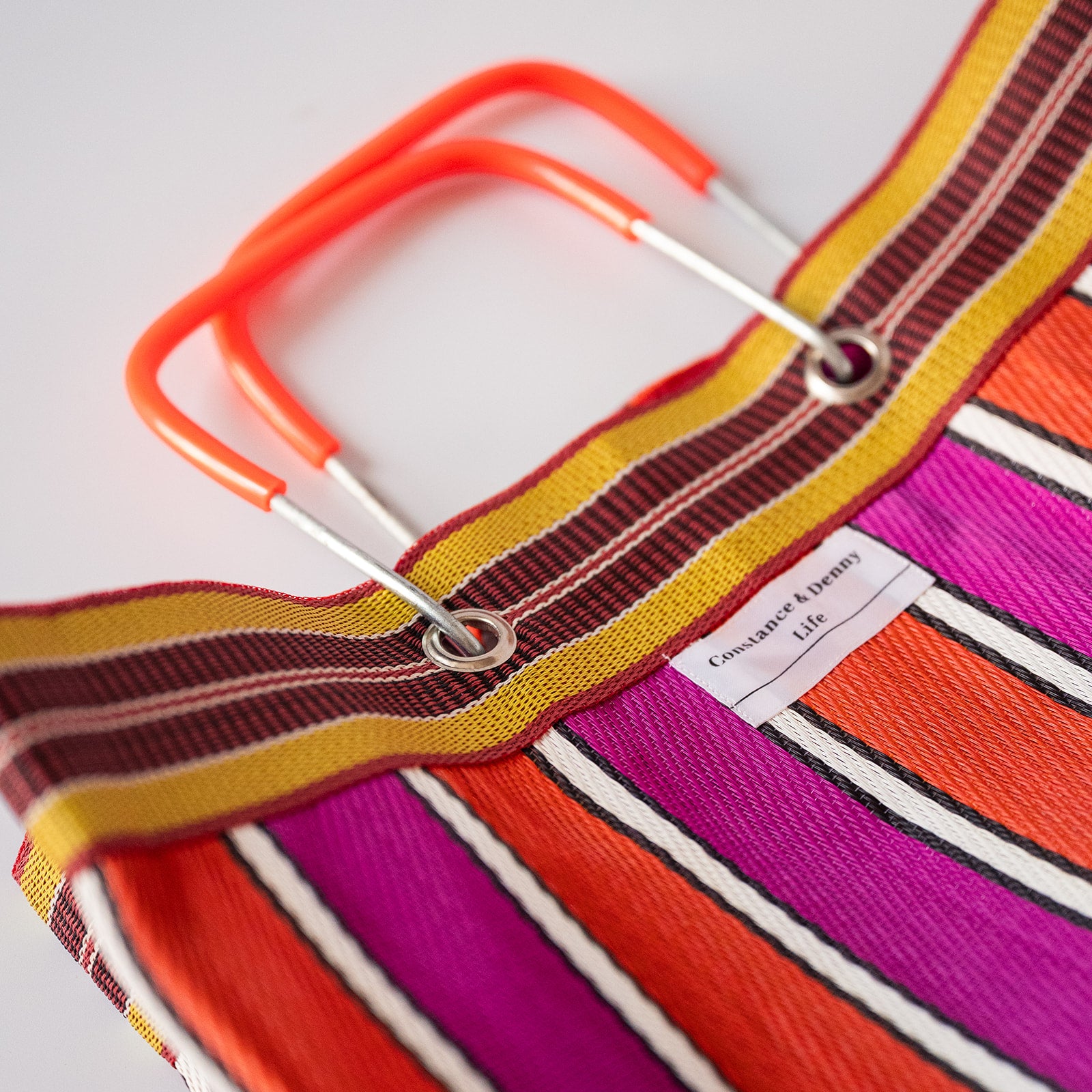 Close-up of a striped tote bag with bright orange handles, featuring bold stripes in orange, magenta, yellow, brown, and white. A small fabric label reads 'Conscious Living'.