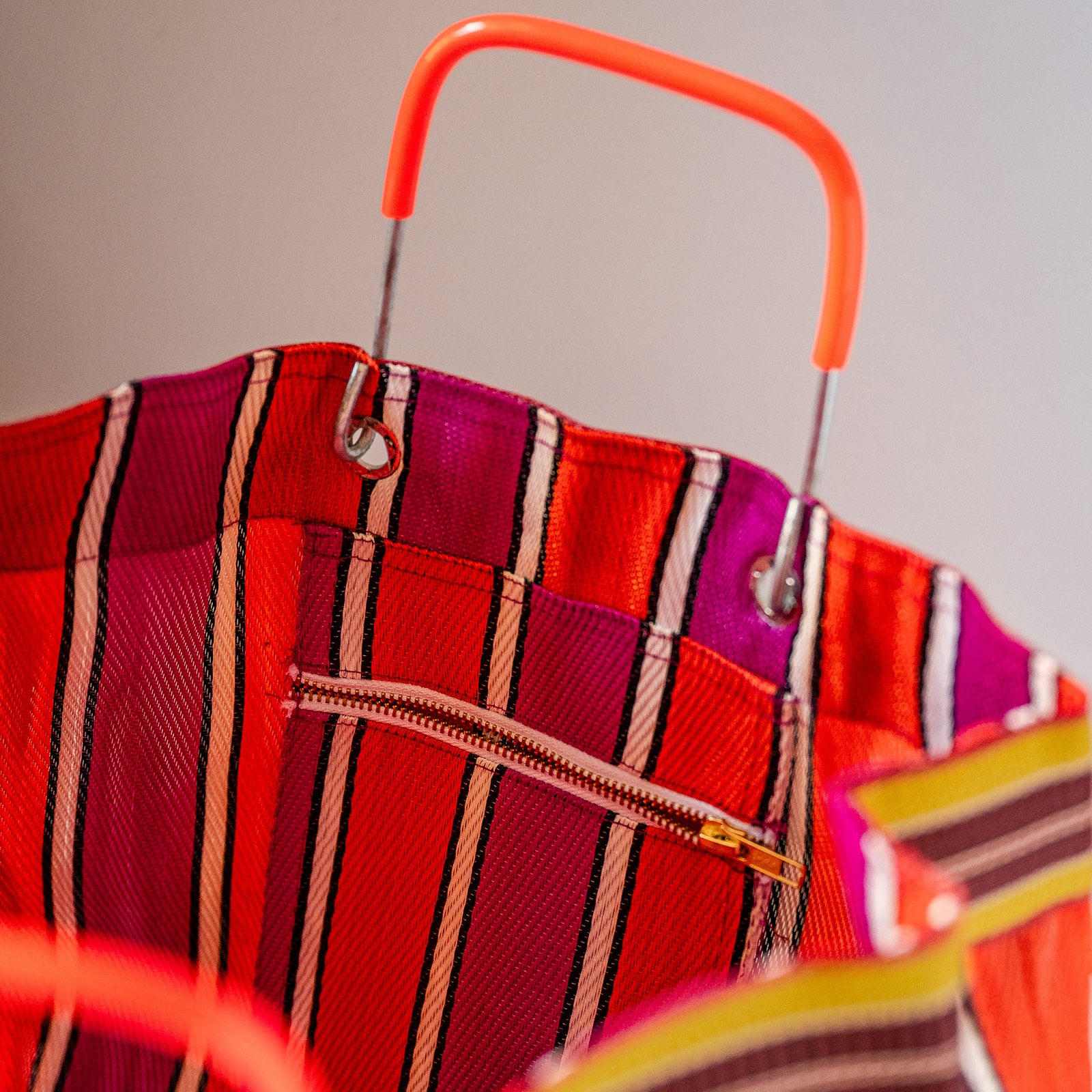 Close-up of a striped tote bag with a bright orange handle, featuring red, pink, and beige vertical stripes and a zippered pocket.