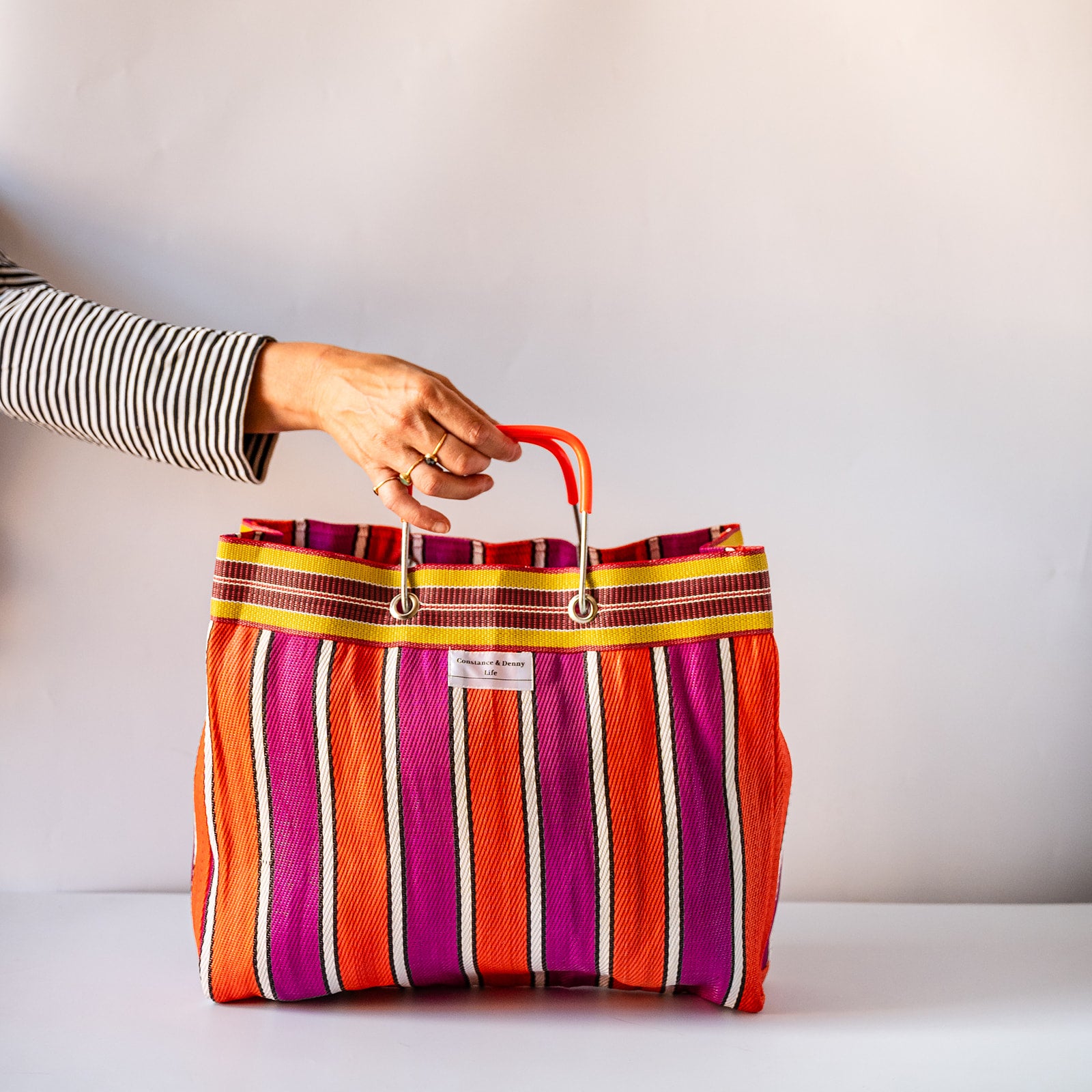 RECYCLED PLASTIC striped tote bag with a bright orange handle, featuring red, pink, and beige vertical stripes and a zippered pocket.