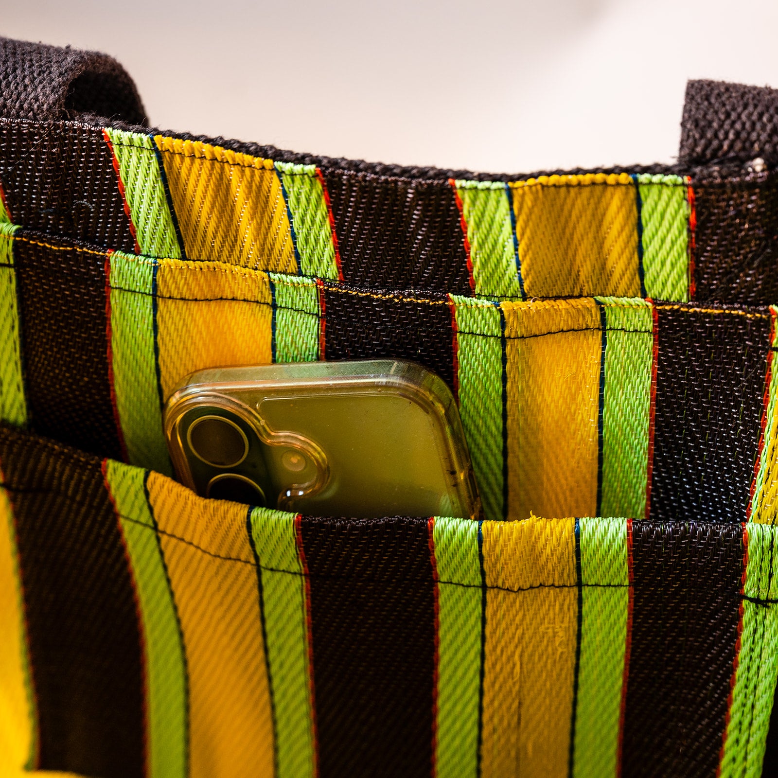 Close-up of a woven tote bag with vertical stripes in black, yellow, and lime green, showing a phone tucked into one of the front pockets.