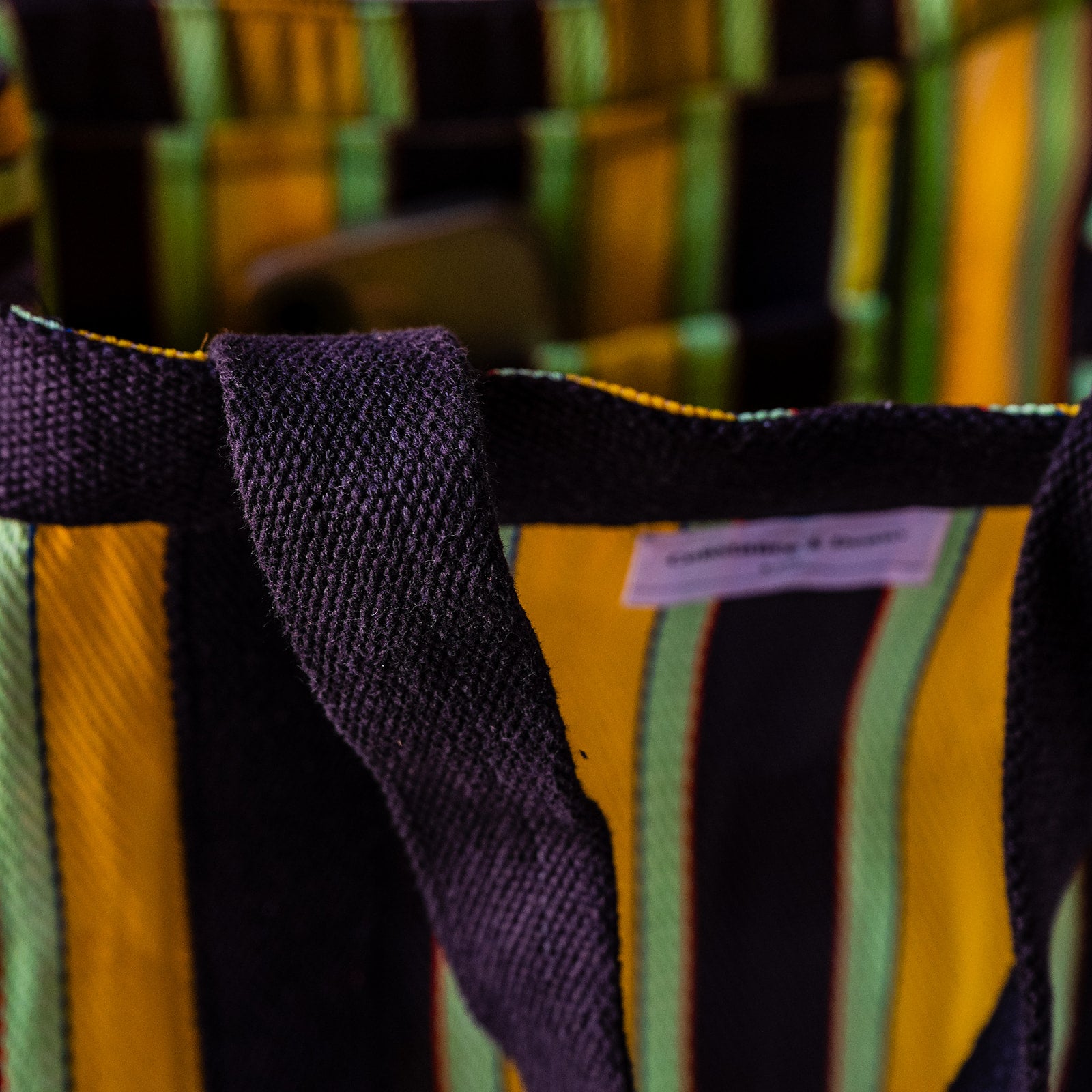 A close-up of a woven tote bag with vertical stripes in yellow, black, and light green, featuring dark purple handles and a small white brand label.