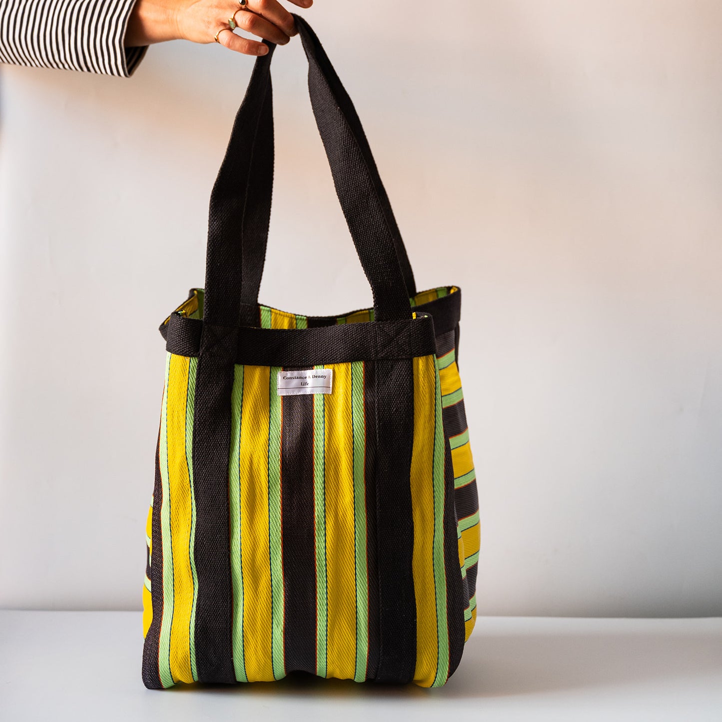 A striped fabric tote bag with yellow, green, and black vertical stripes, held by a hand wearing a black and white striped shirt against a plain white background.