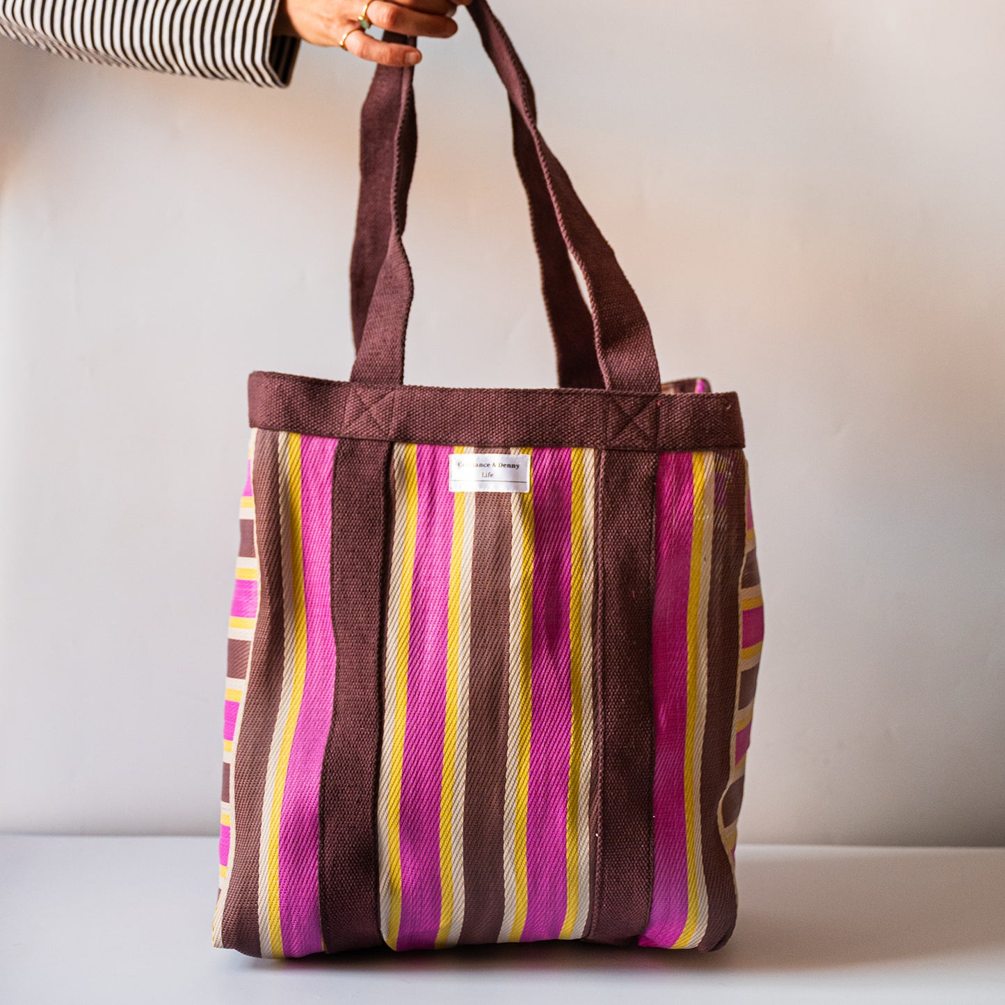 A striped canvas tote bag with dark brown handles and a vertical stripe pattern in brown, pink, yellow, and white. A hand wearing a striped long-sleeve shirt holds the top of the bag against a plain light gray background.