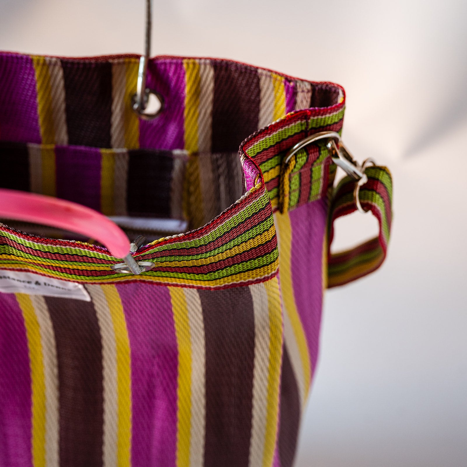Close-up of a woven tote bag with vertical purple, magenta, yellow, brown, and cream stripes, featuring a pink handle and metal hardware.