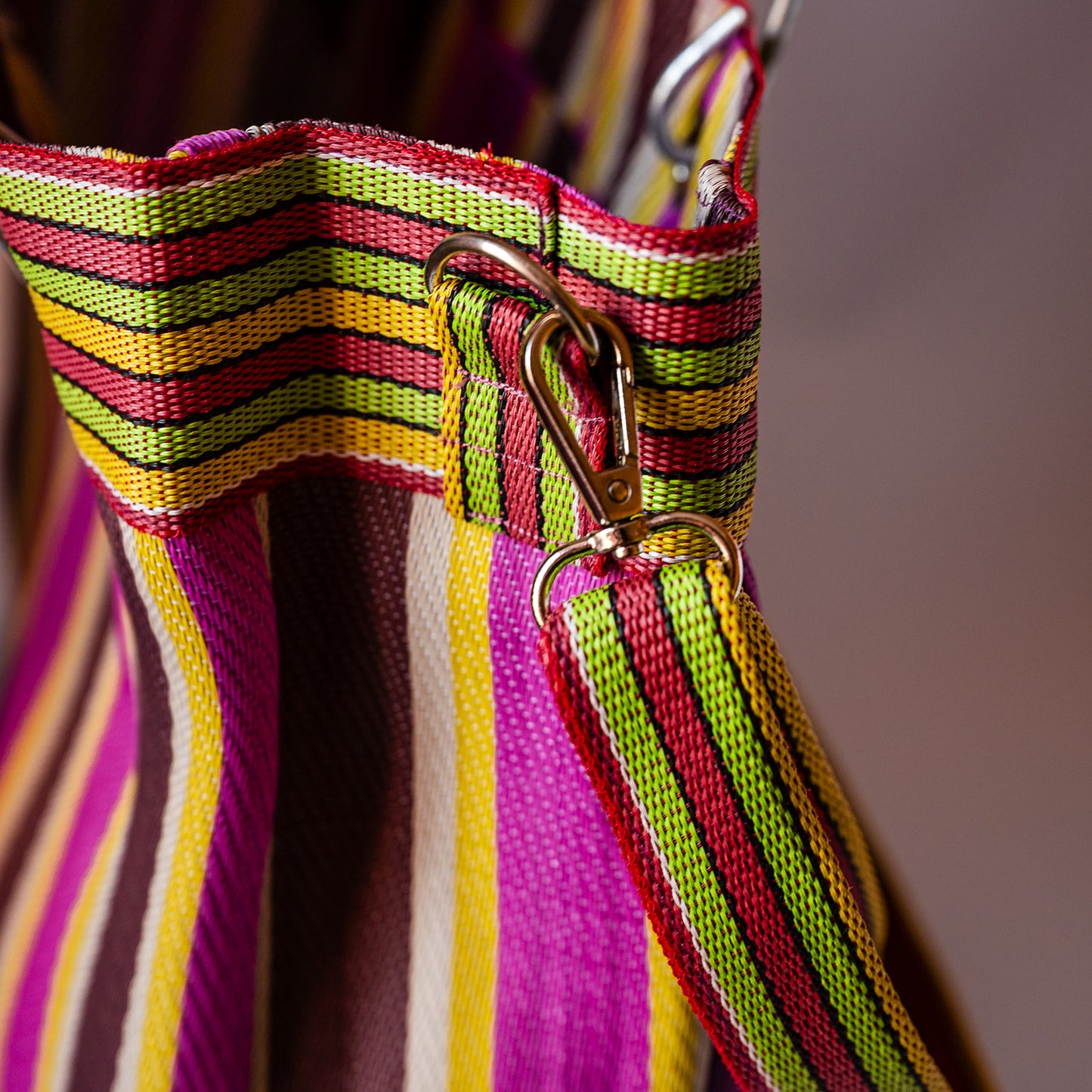 Close-up detail of a striped fabric bag with a woven strap featuring pink, green, yellow, and white stripes, attached to a gold-tone metal swivel clasp.