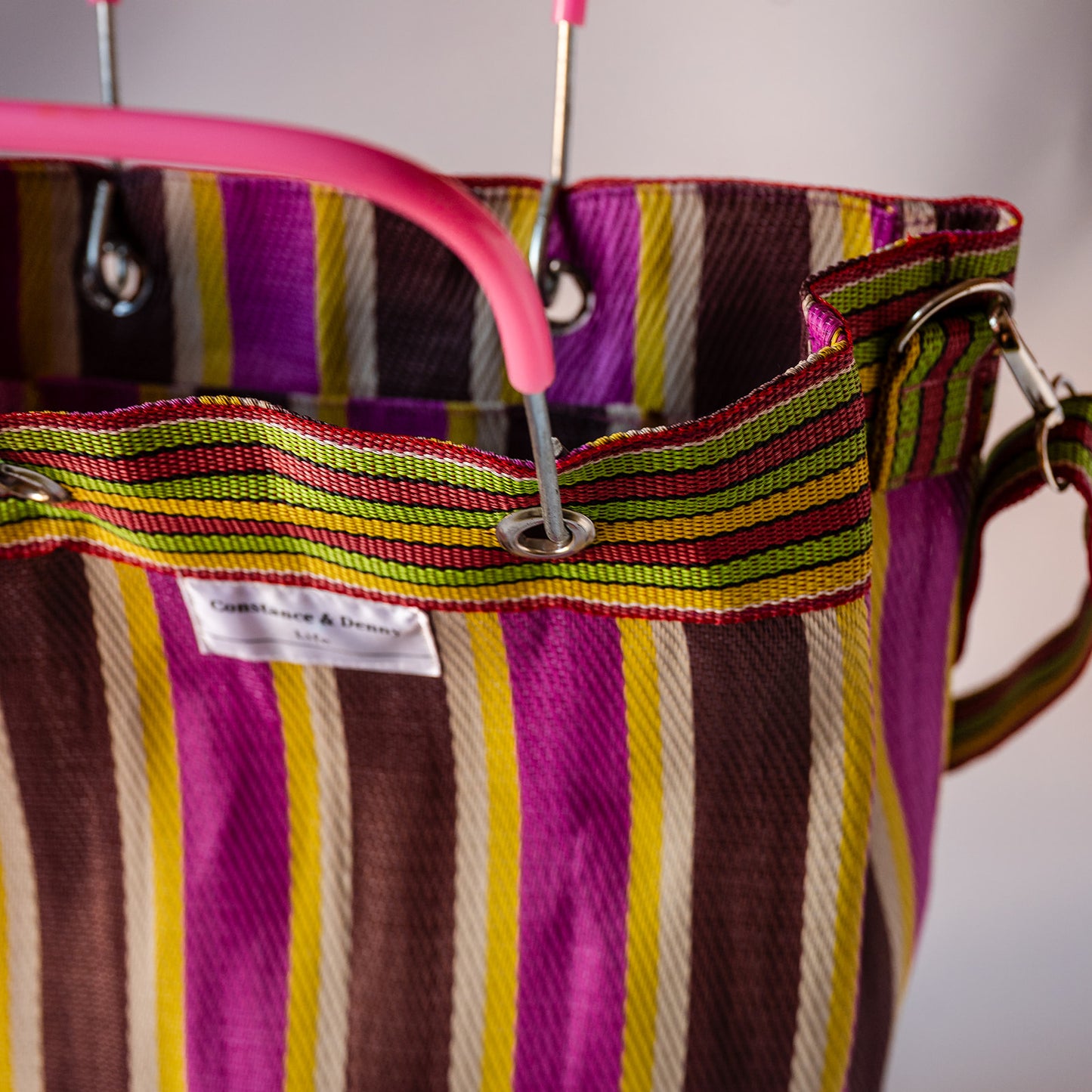 Close-up of a striped fabric tote bag with a pink handle, featuring vertical stripes in purple, brown, yellow, green, and cream. A white fabric label is visible on the front.