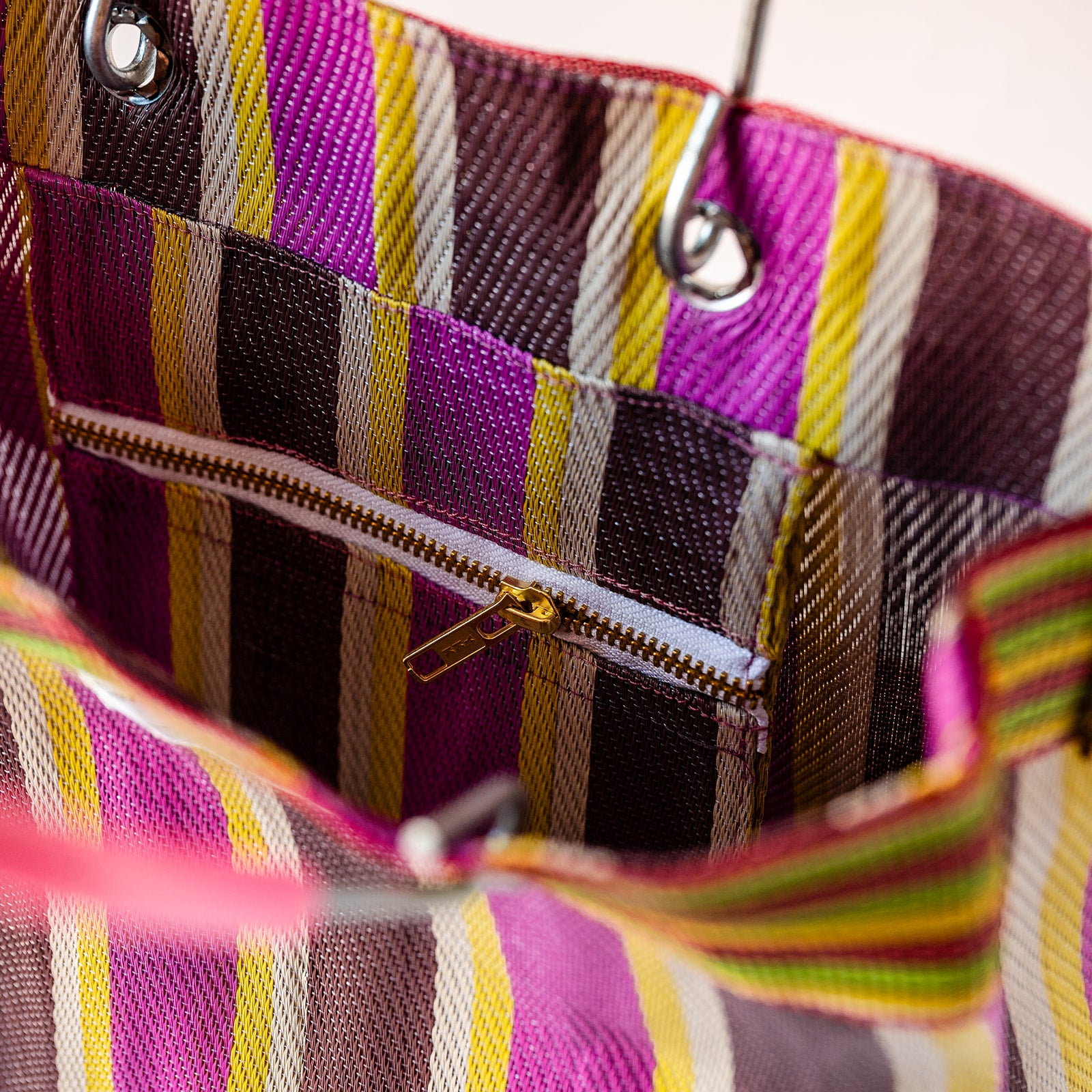 Close-up of the interior of a striped tote bag, showing a zippered pocket with a gold zipper, a woven fabric lining in pink, purple, brown, yellow, and white stripes, and metal grommets.