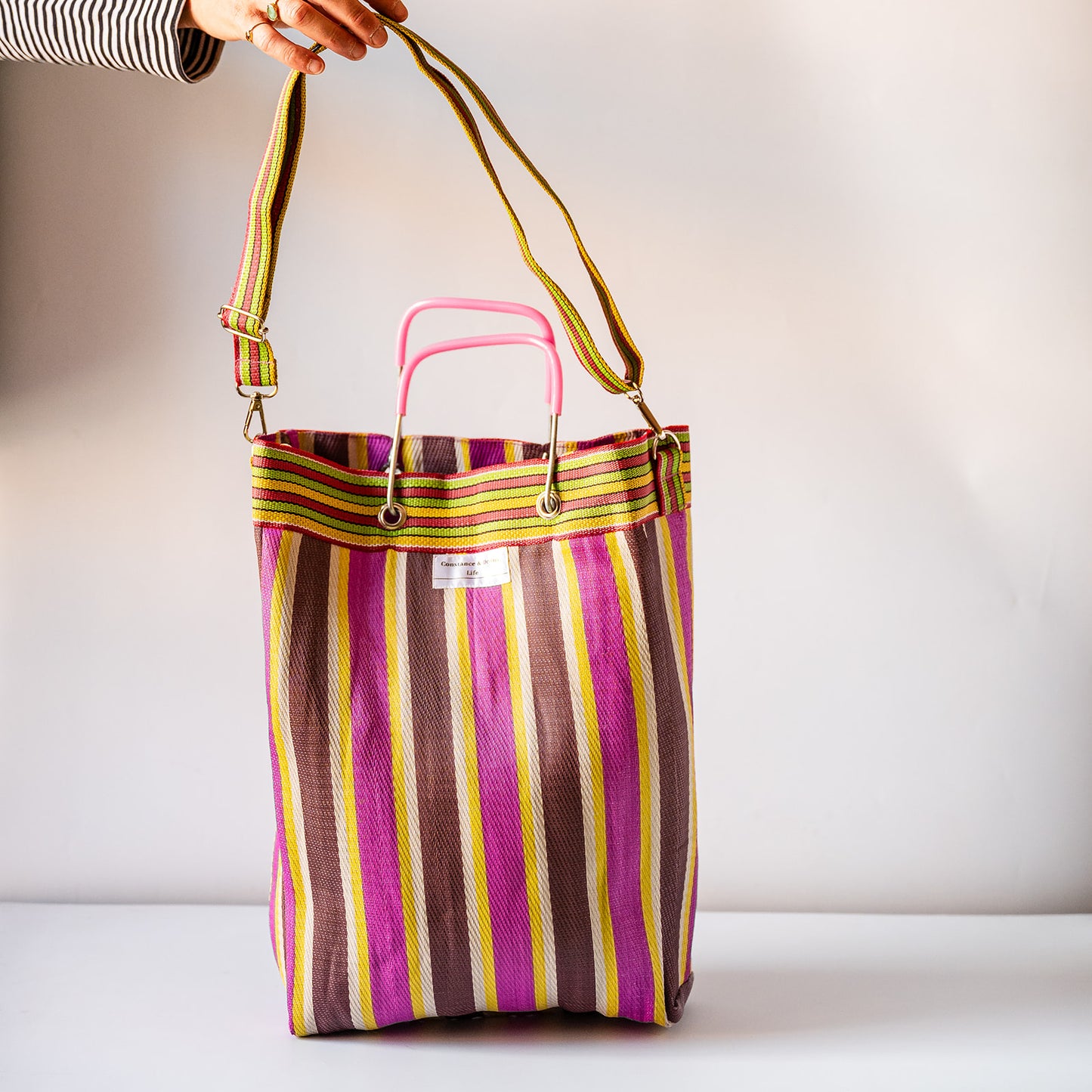 A striped tote bag with vertical pink, purple, brown, yellow, and green stripes, featuring a pink top handle and a long adjustable shoulder strap held by a hand.