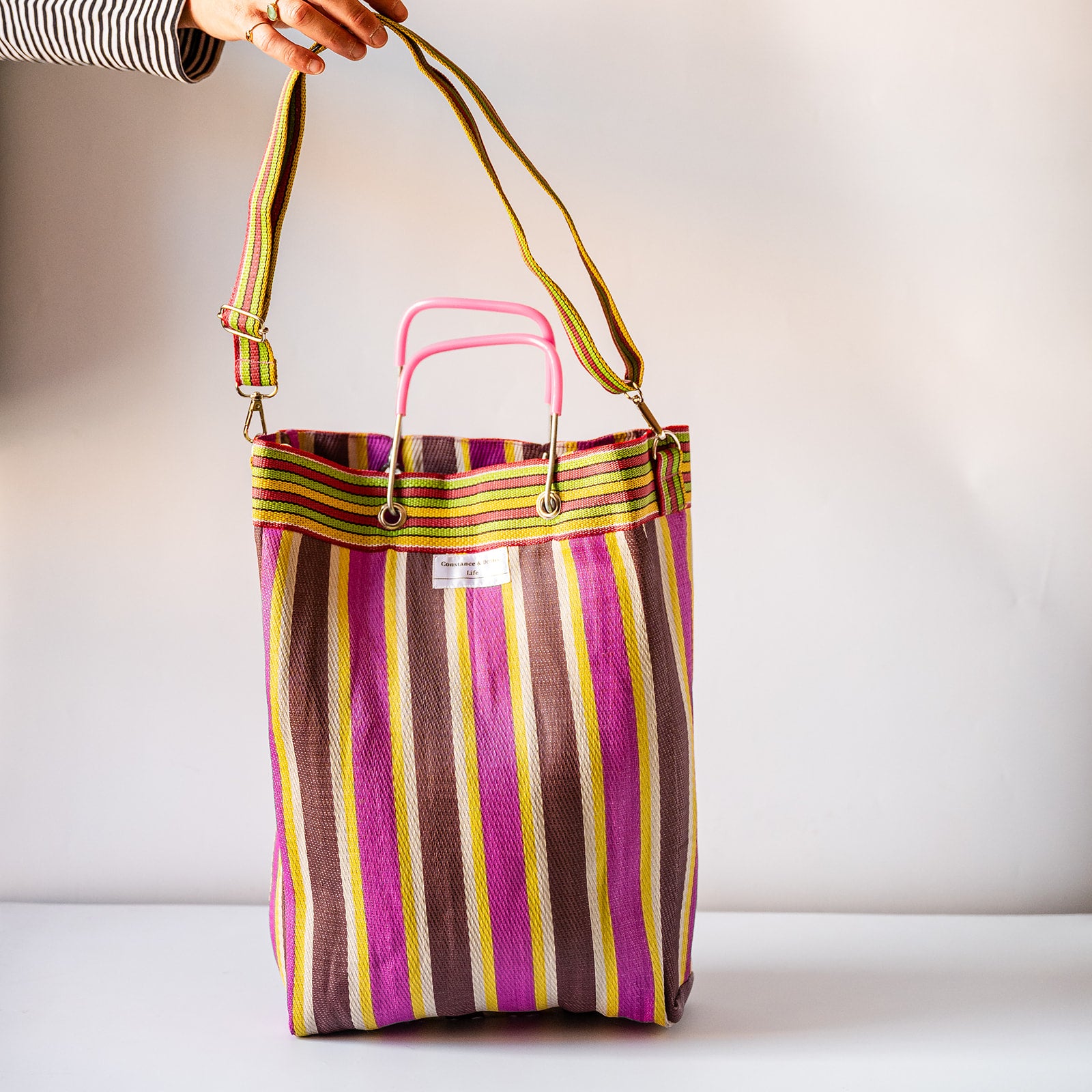 A striped tote bag with vertical pink, purple, brown, yellow, and green stripes, featuring a pink top handle and a long adjustable shoulder strap held by a hand.