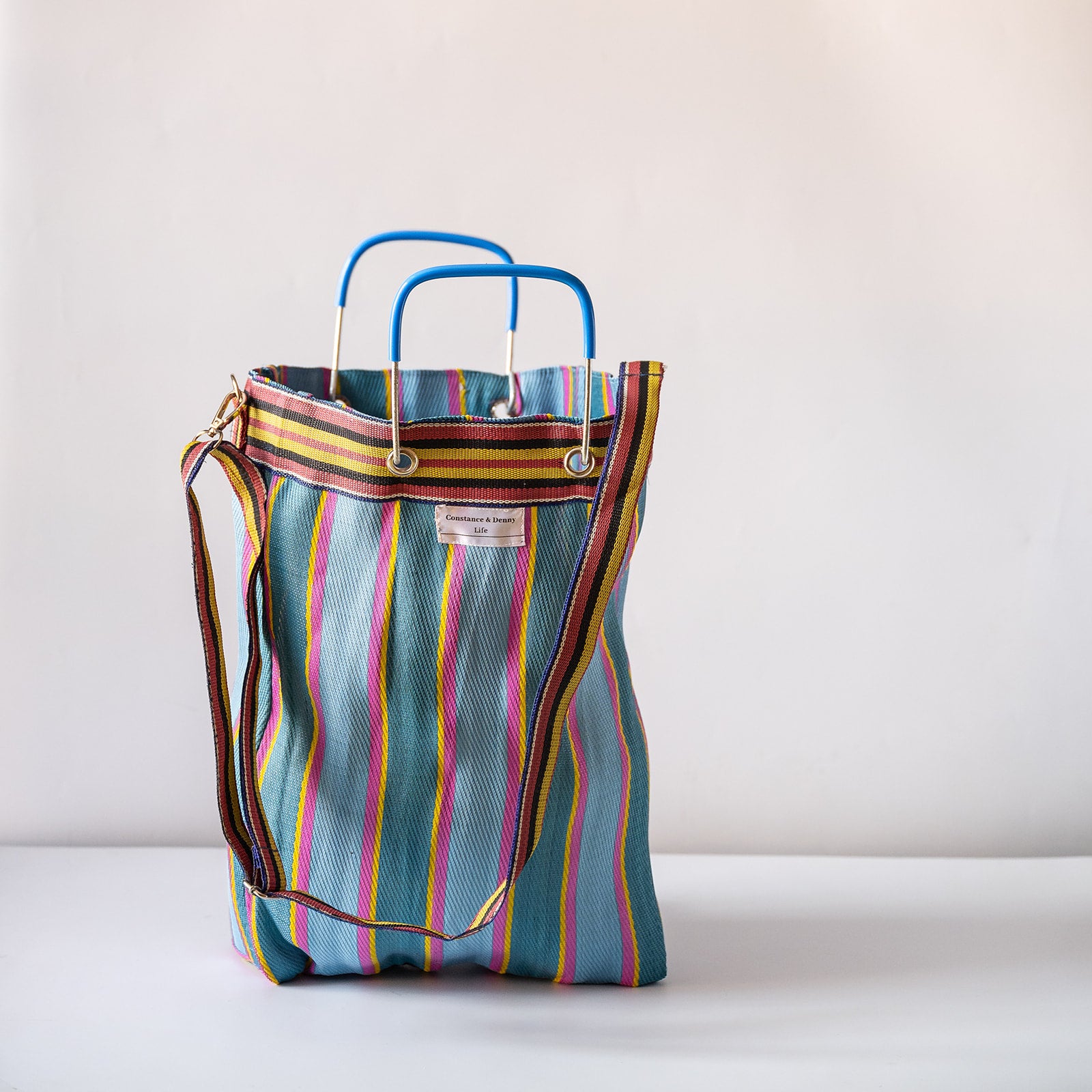 A striped canvas tote bag with a blue top handle and shoulder strap, featuring vertical stripes in teal, yellow, pink, and brown on a plain light background.