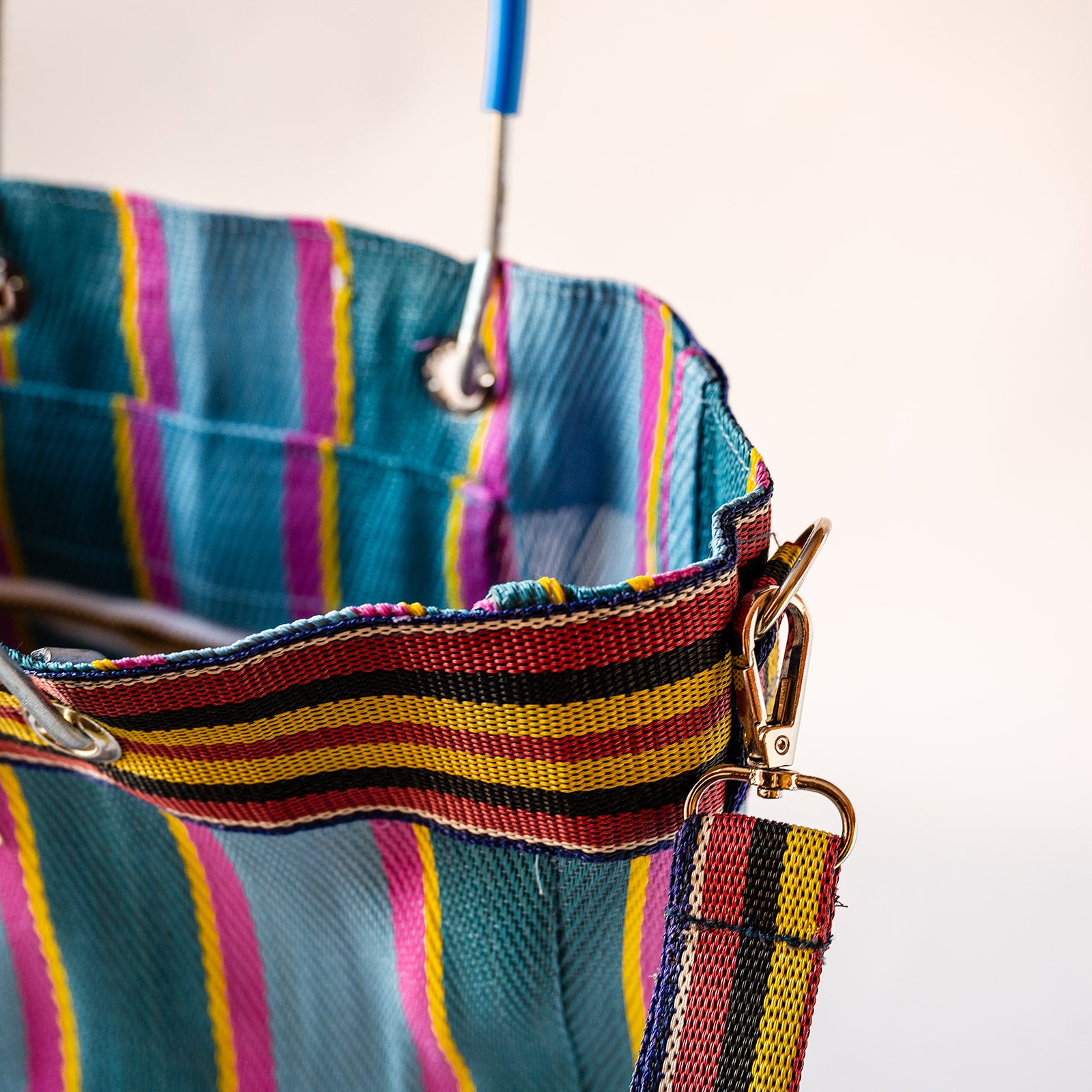 Close-up of a colorful striped fabric bag or tote with a woven strap, metal grommets, and gold-tone hardware against a plain light background.