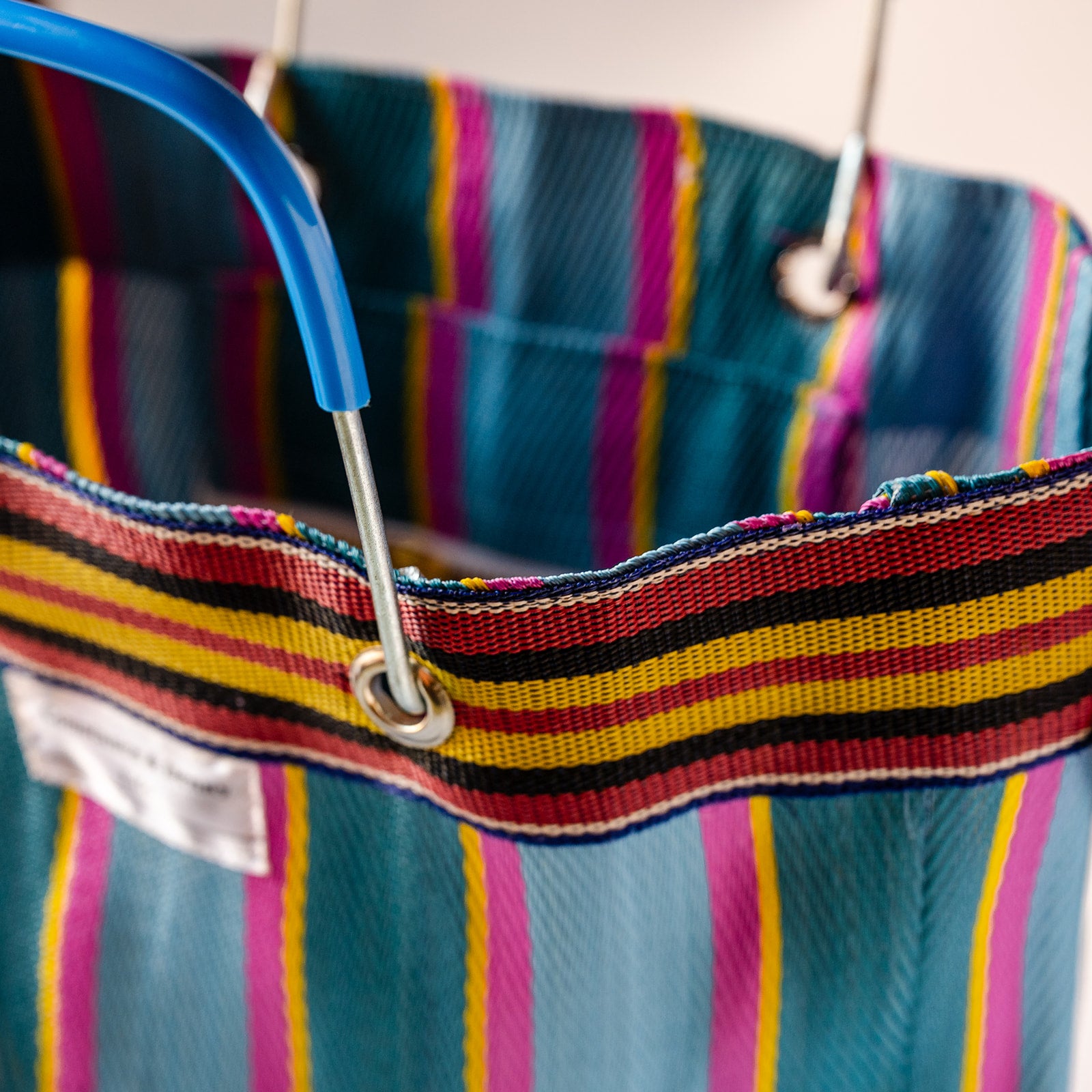Close-up of a striped fabric tote bag with a blue plastic handle and metal grommets, showing teal, pink, yellow, red, and black woven stripes.