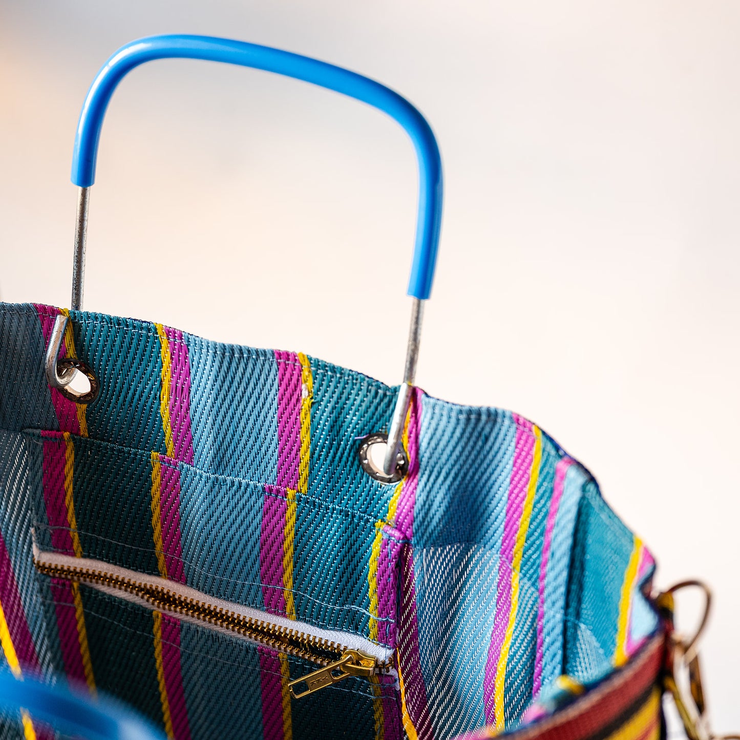 Close-up of a colorful striped fabric tote bag with a bright blue top handle and a zippered pocket, photographed against a soft light background.