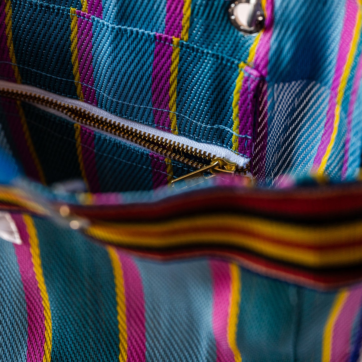 Close-up of a striped fabric product with vertical magenta, yellow, and teal stripes, showing a gold zipper and metal snap detail.