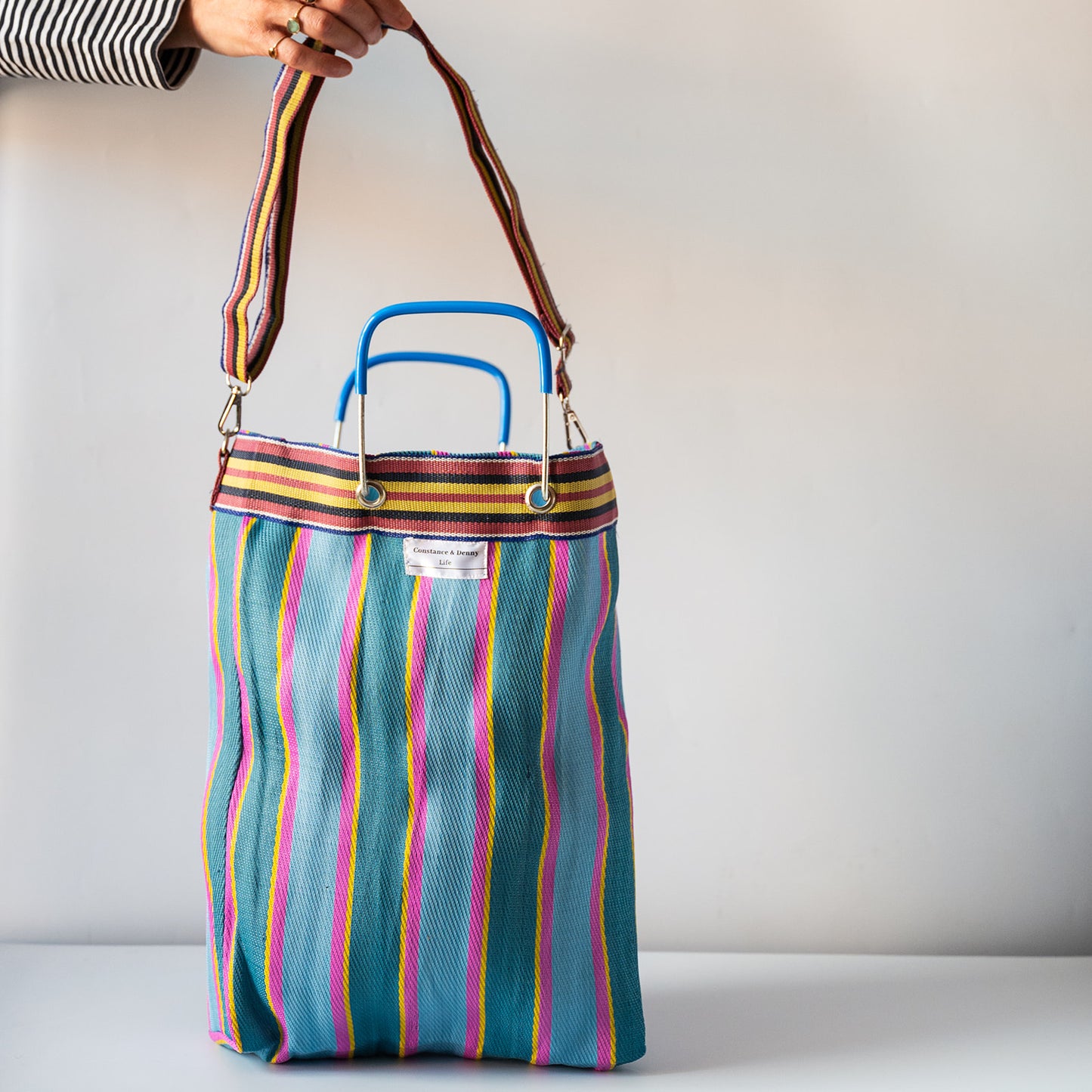 A striped tote bag with a teal base and vertical pink, yellow, orange, and white stripes, featuring blue top handles and a brown shoulder strap, held by a hand against a plain light gray background.
