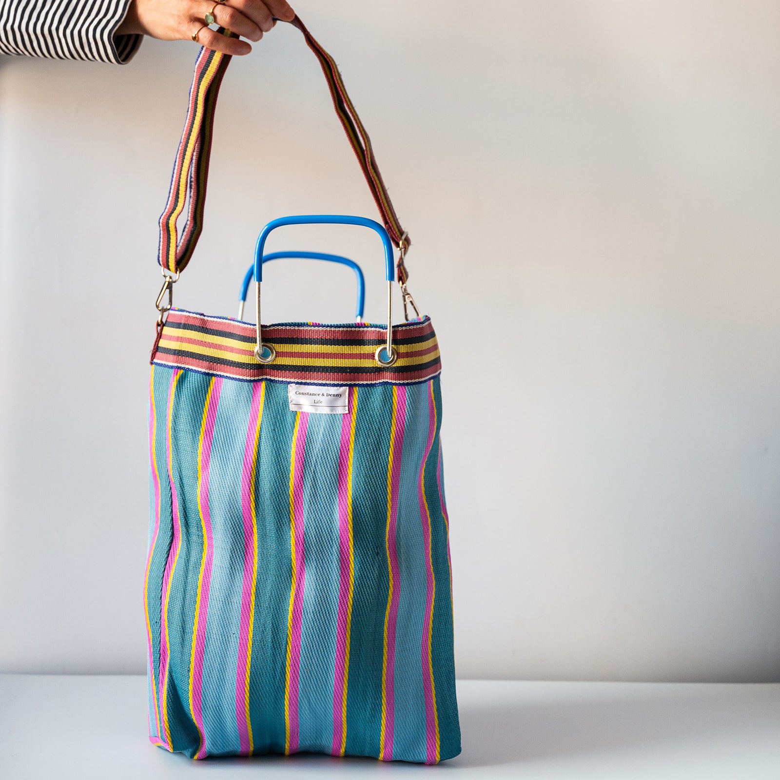 A striped tote bag with a teal base and vertical pink, yellow, orange, and white stripes, featuring blue top handles and a brown shoulder strap, held by a hand against a plain light gray background.