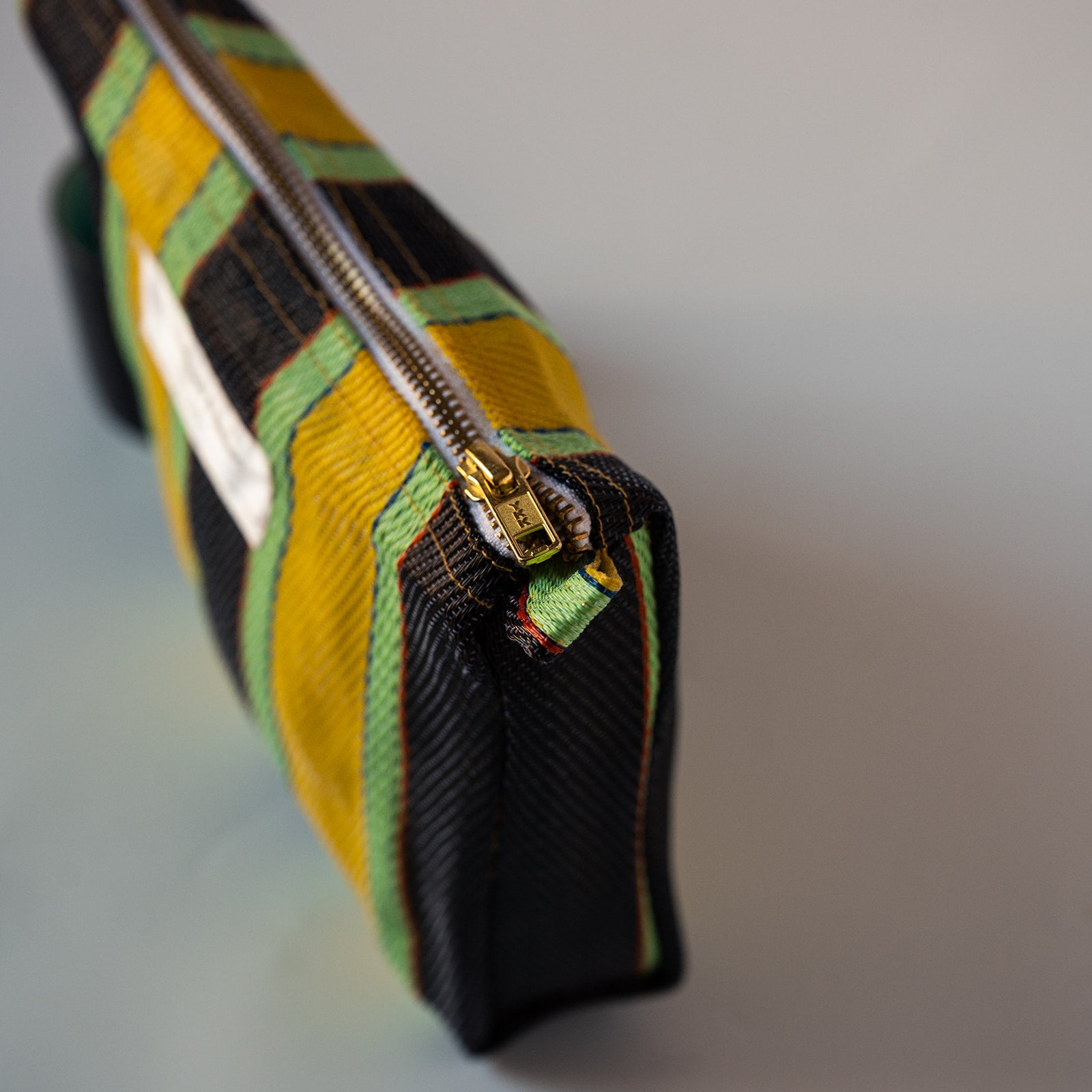 A close-up of a woven fabric zip pouch with a bold plaid pattern in yellow, black, and green, featuring a gold-tone metal zipper.