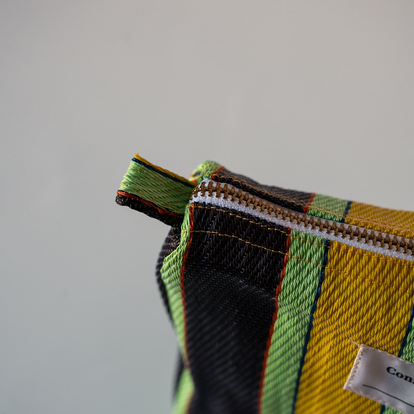 Close-up of a woven zippered pouch corner showing a striped plaid pattern in yellow, green, black, and orange, with a white fabric brand tag partially visible.
