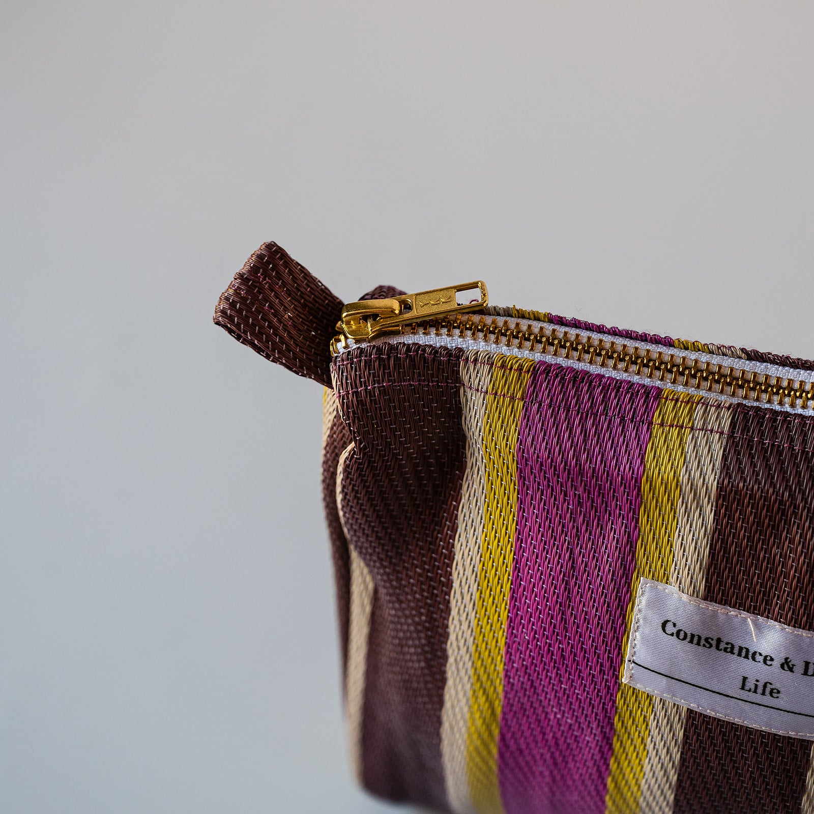 Close-up of a striped fabric zip pouch with a gold zipper and a white Constance & Life label, set against a plain light gray background.
