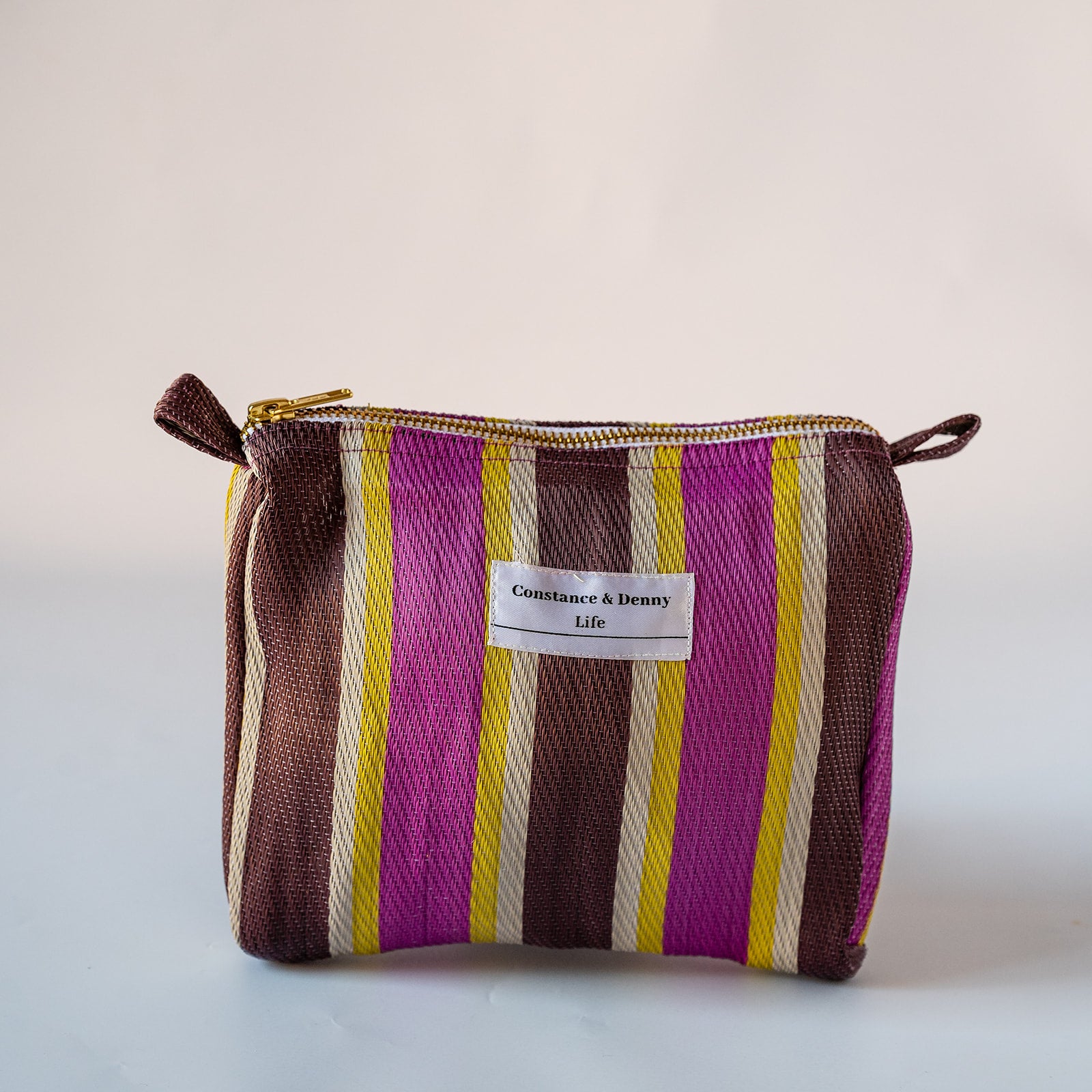 A small zippered cosmetic pouch with a woven striped pattern in brown, magenta, yellow, and cream, featuring a white label that reads 'Countance & Benny Life'.