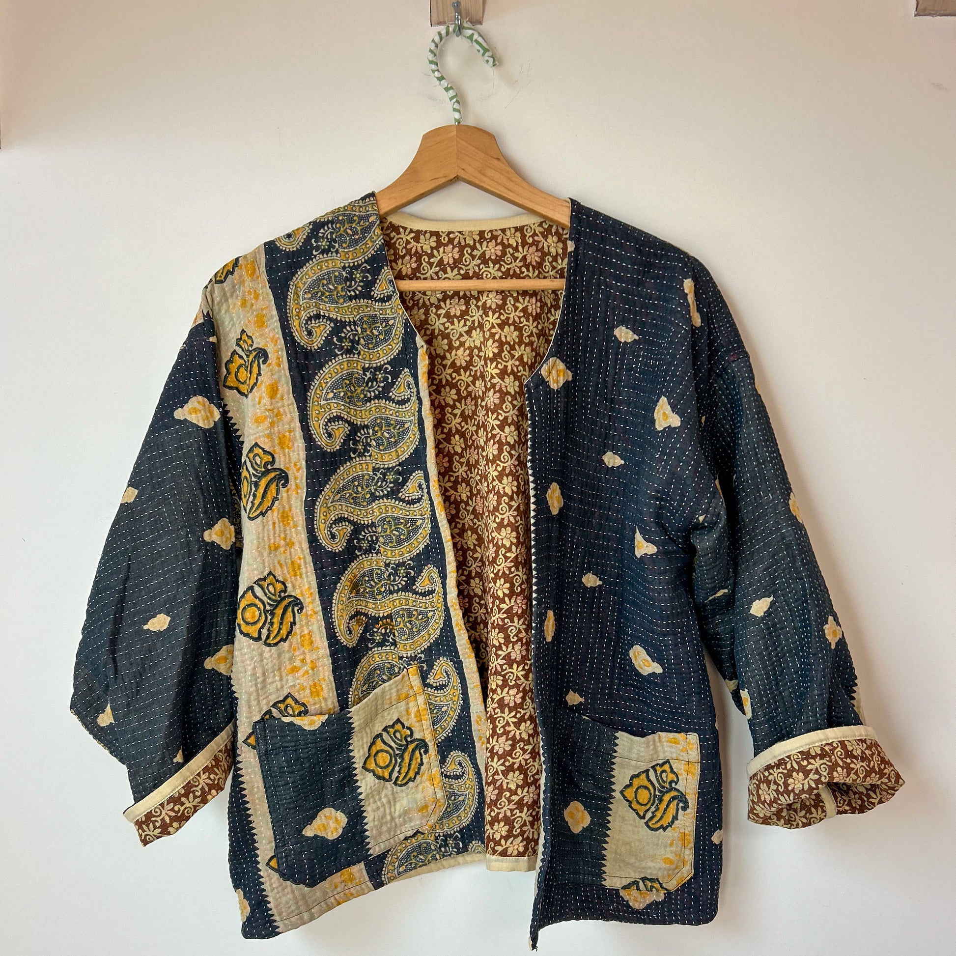 Traditional patterned kantha jacket on a hanger against a white wall. Vintage-kantha-jacket-handmade-unique-37-paisley-pattern