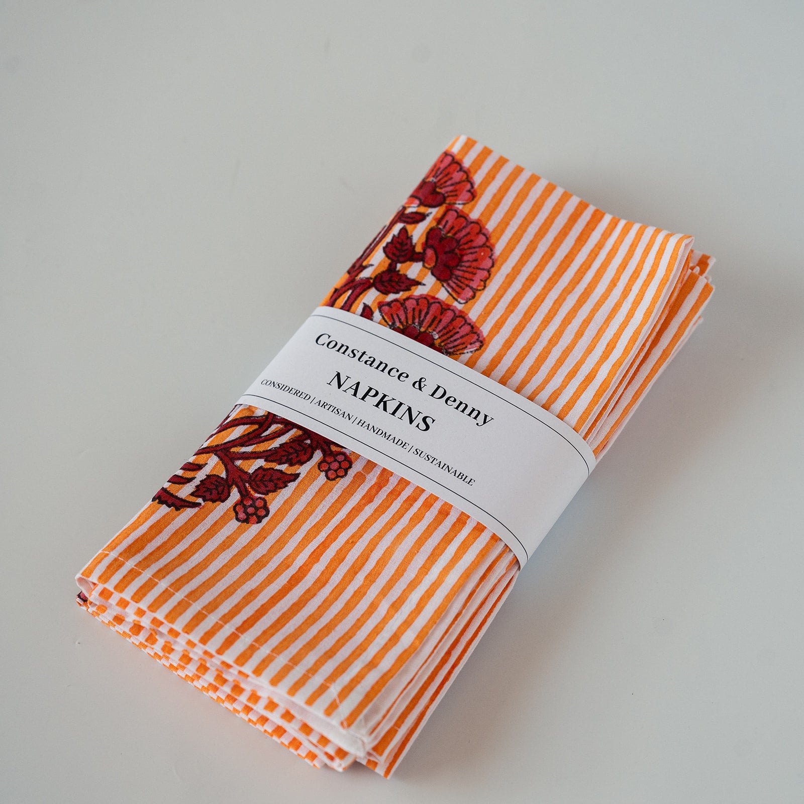 A folded set of Constance & Denny napkins with orange and white stripes and a red floral pattern, wrapped in a white paper band.