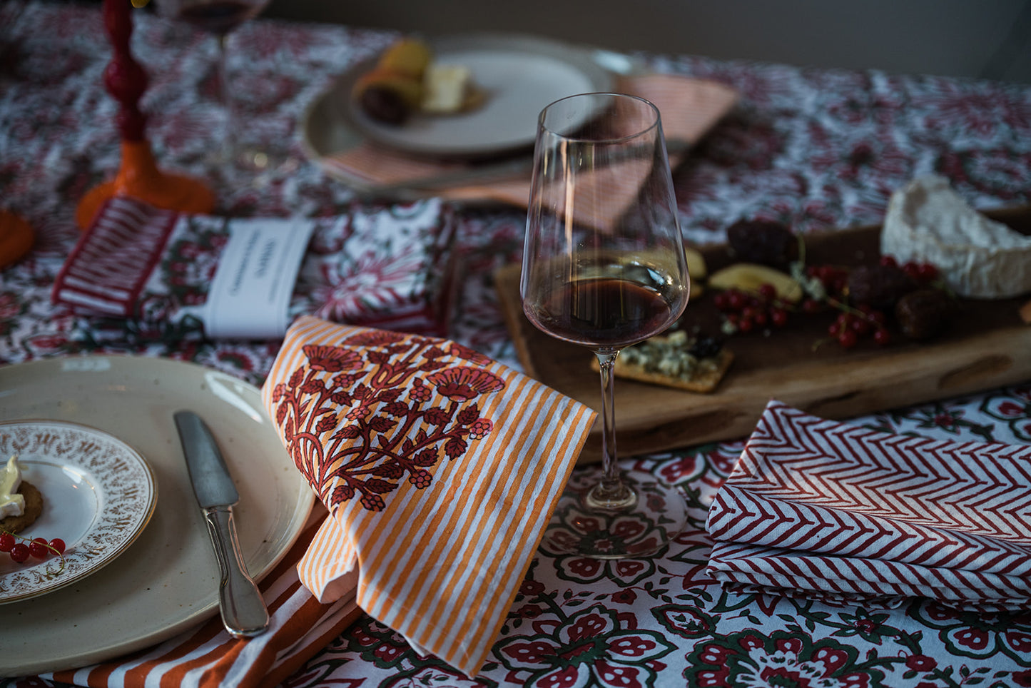 A styled dining table setting with a patterned tablecloth, a wine glass of red wine, a cheese board with crackers and fruit, plates, and folded striped and patterned napkins.