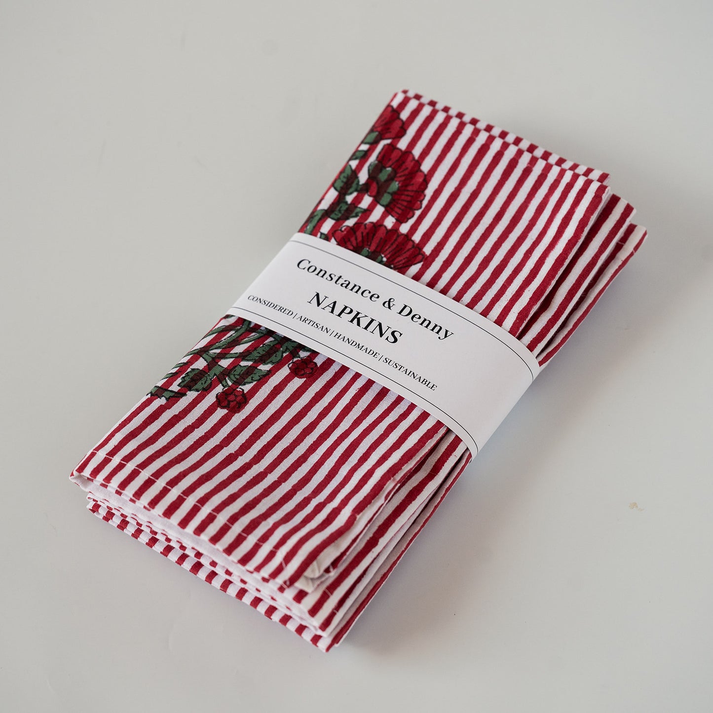 A stack of folded Constance & Denny napkins with red and white stripes and embroidered red roses, wrapped in a white paper band label.