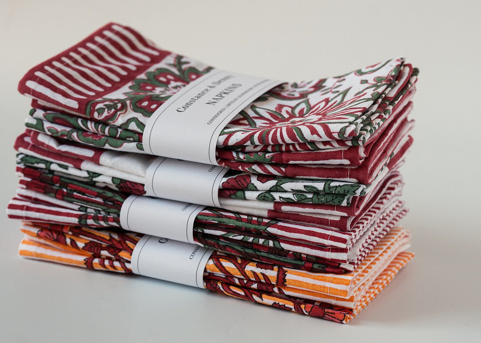 A stack of folded cotton kitchen towels with various holiday and autumn patterns, including red and white stripes, red and green floral motifs, and orange and brown leaf designs, wrapped with white paper bands.