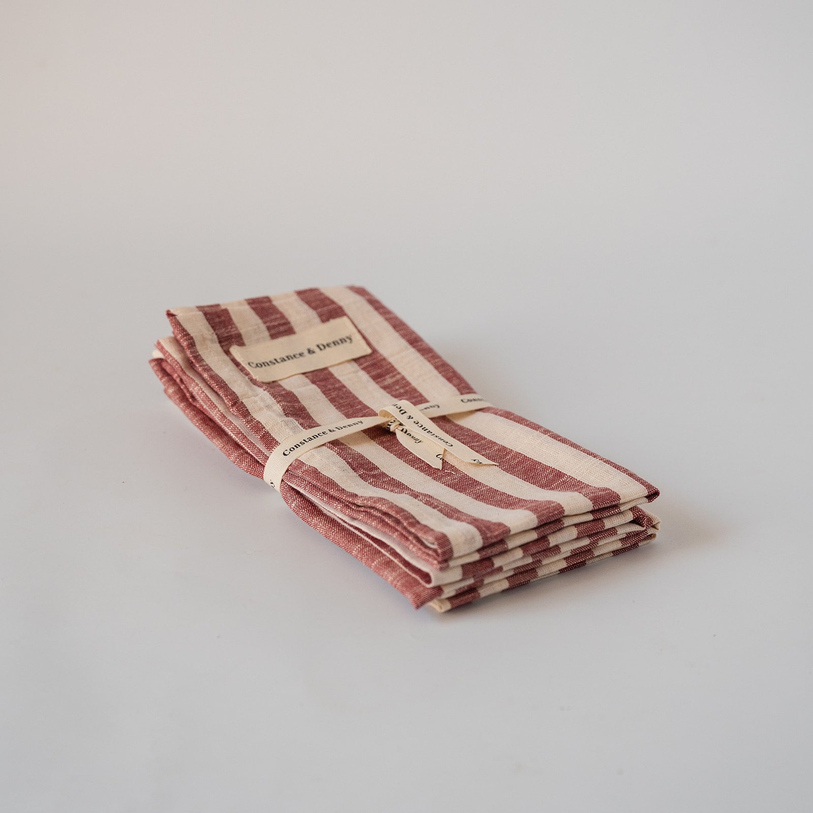 A folded stack of striped linen napkins in muted terracotta red and cream, tied with a paper band and ribbon, on a light gray background.