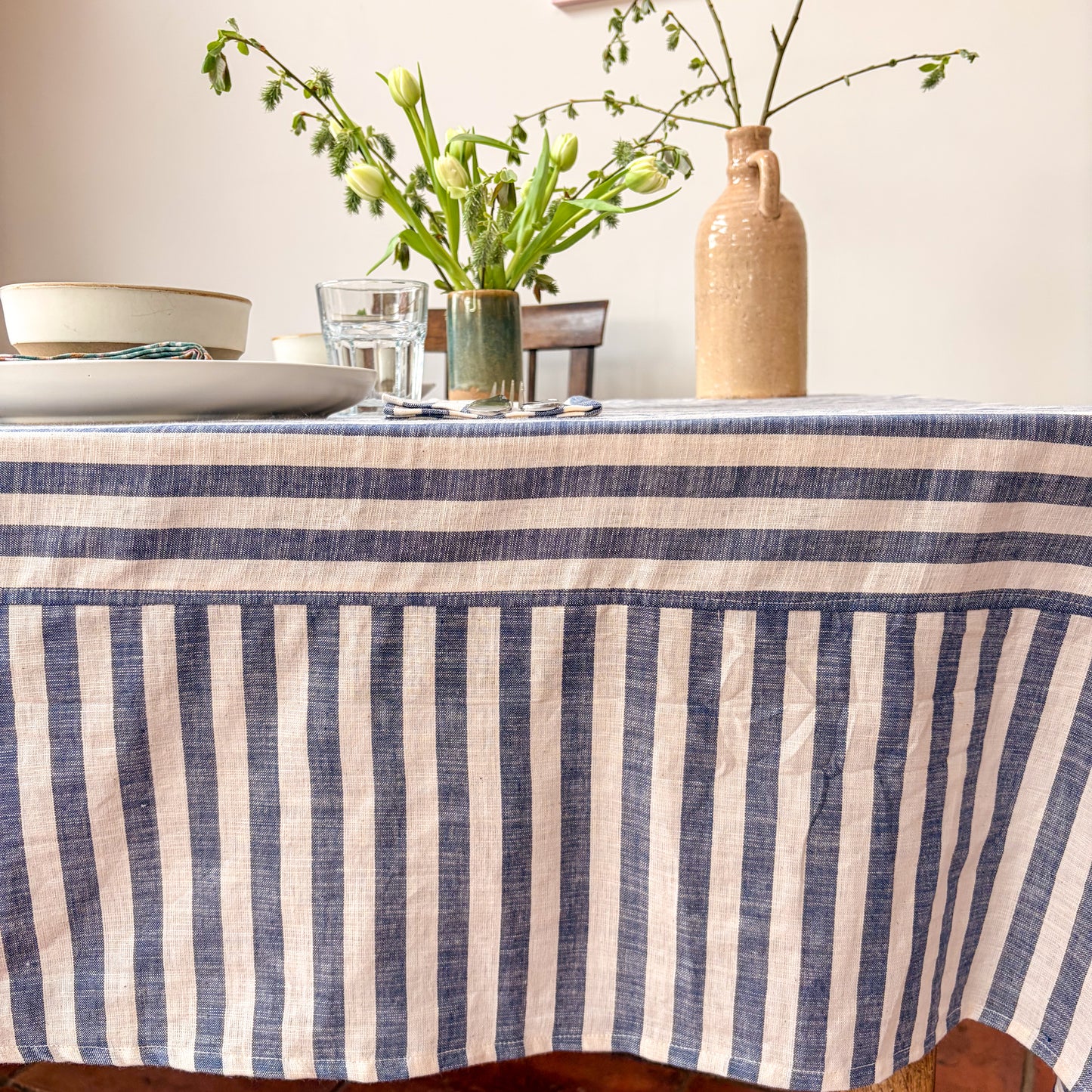 Dining table with a blue and white striped tablecloth, glasses, plates, and a vase with flowers.