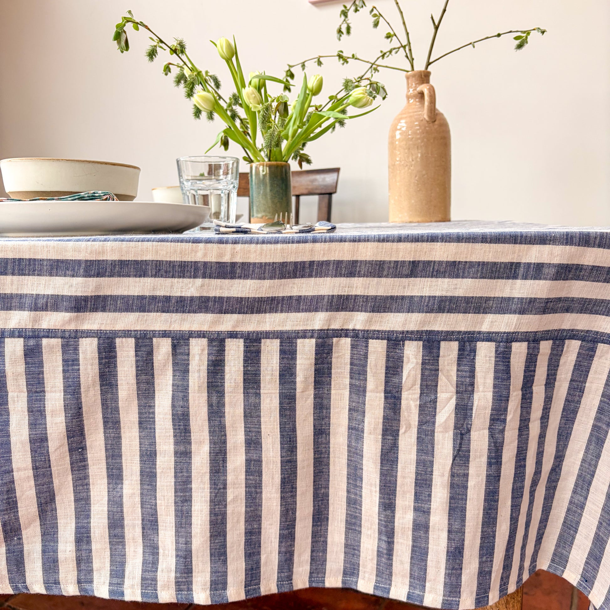 Dining table with a blue and white striped tablecloth, glasses, plates, and a vase with flowers.