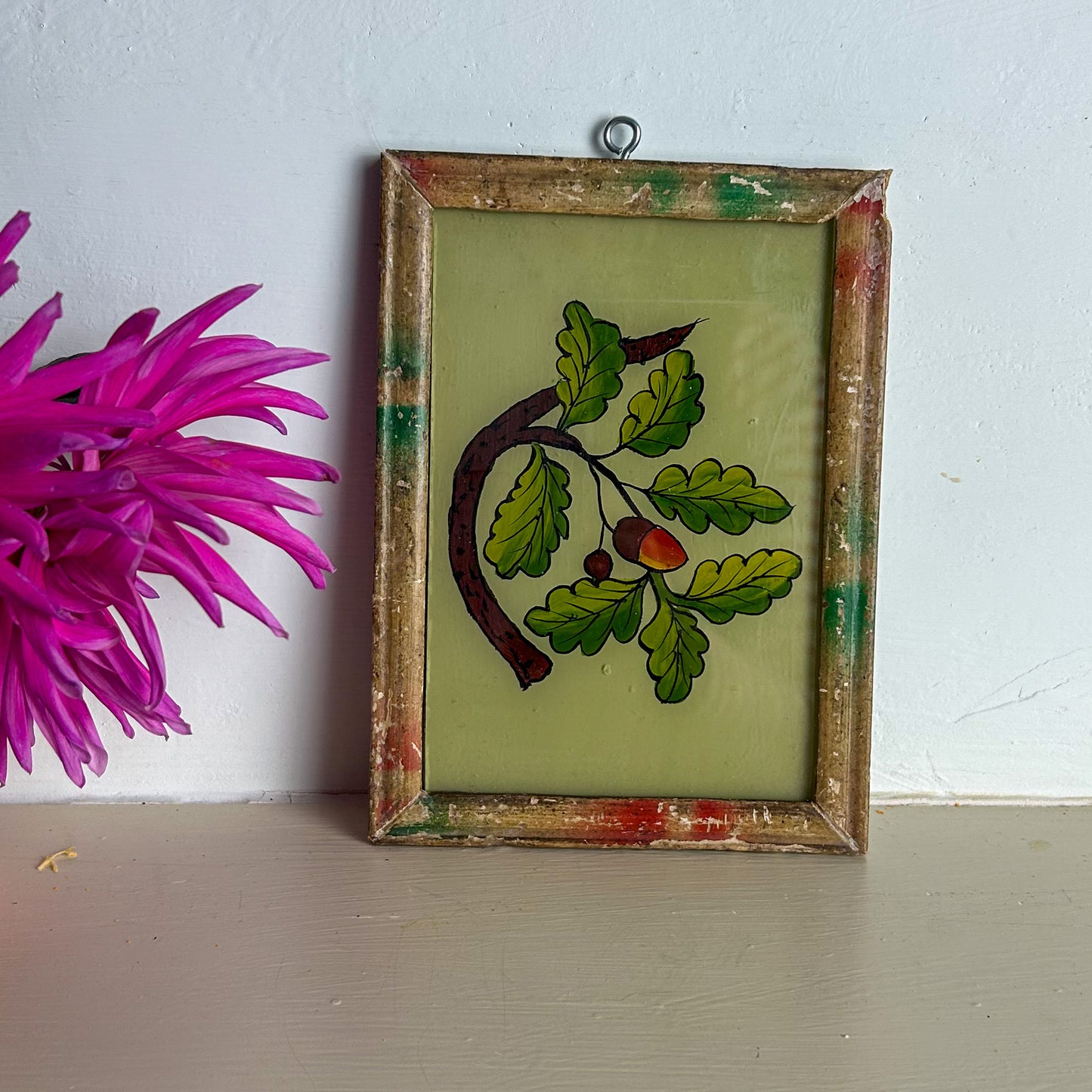 Framed stained glass artwork with leaves and berries next to a pink flower on a white wall.