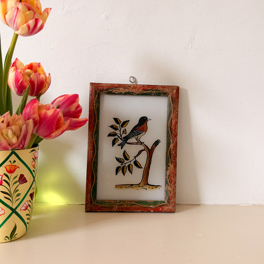 Framed artwork of a bird on a branch next to a vase with tulips on a white surface.