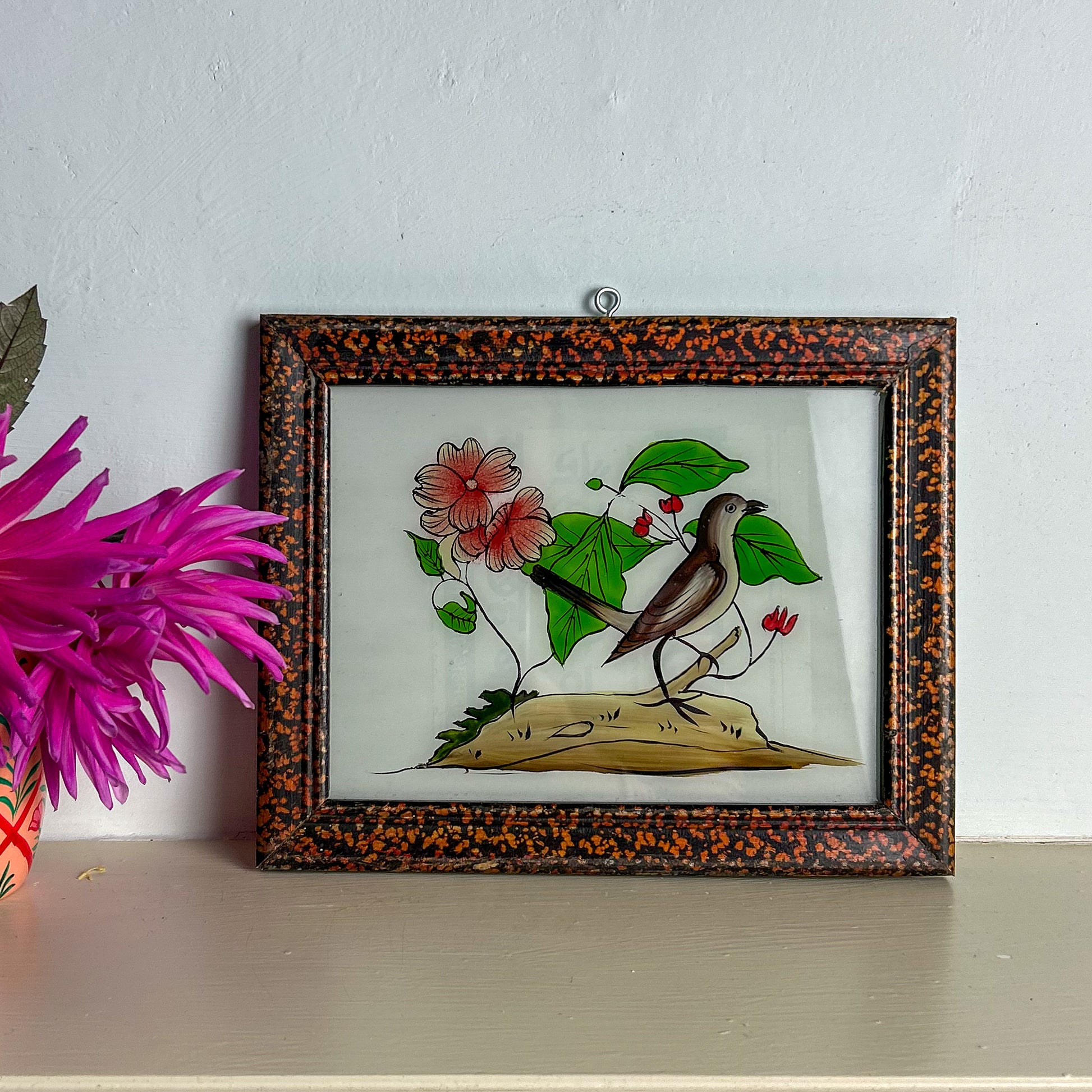Framed artwork of a bird and flowers on a white surface with a pink flower in the foreground.