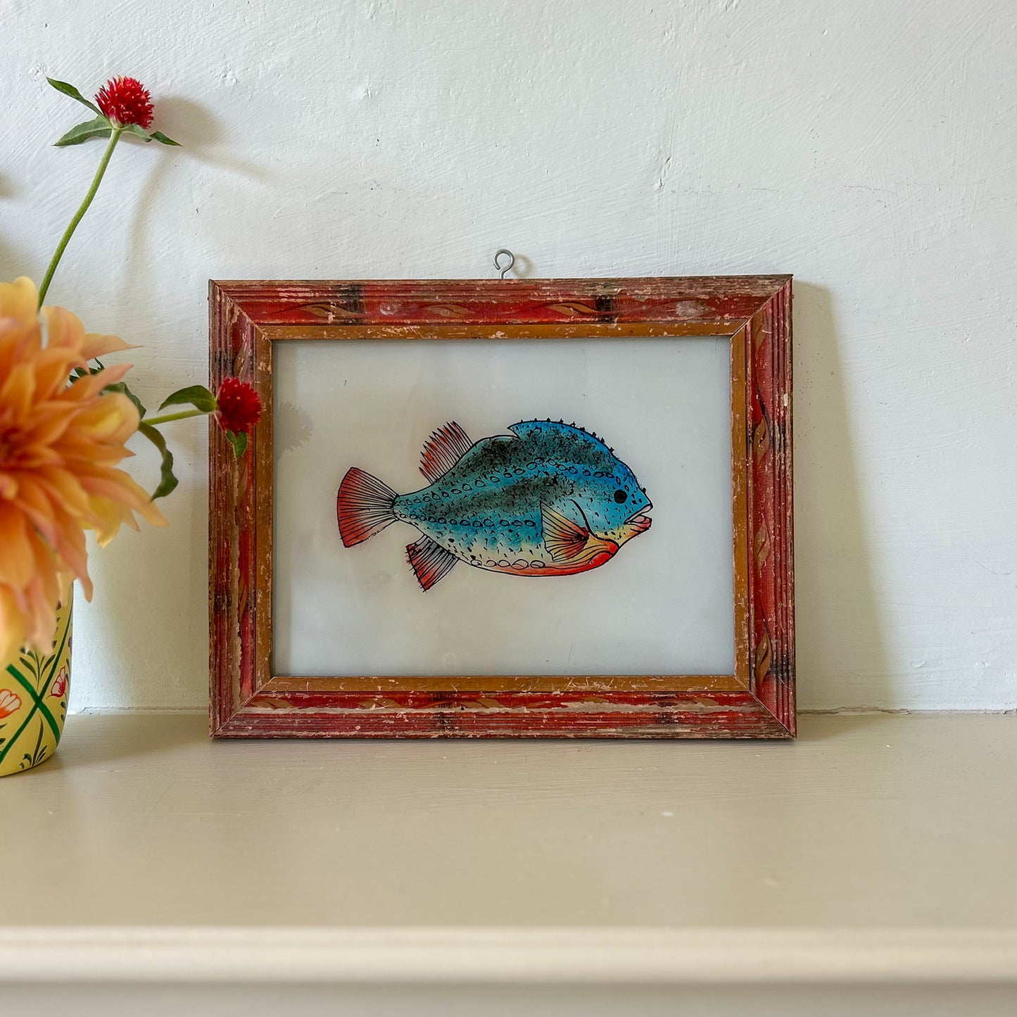 Framed artwork of a blue fish on a white surface with flowers. constance-and-denny-indian-reverse-glass-painting-boxed-packaging-hand-painted-artisan-art-blue-fish