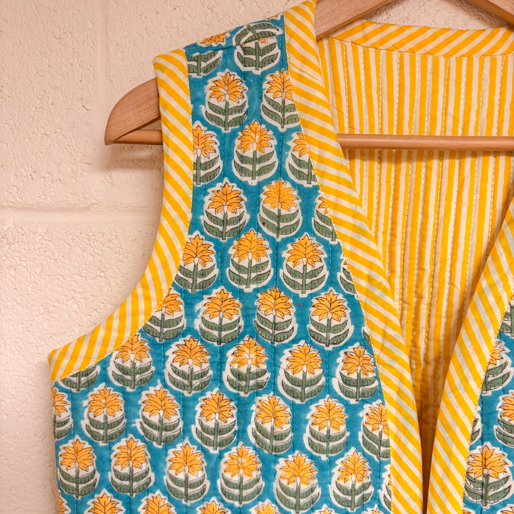 Traditional Indian saree with floral pattern and yellow stripes on a wooden hanger against a light pink wall.