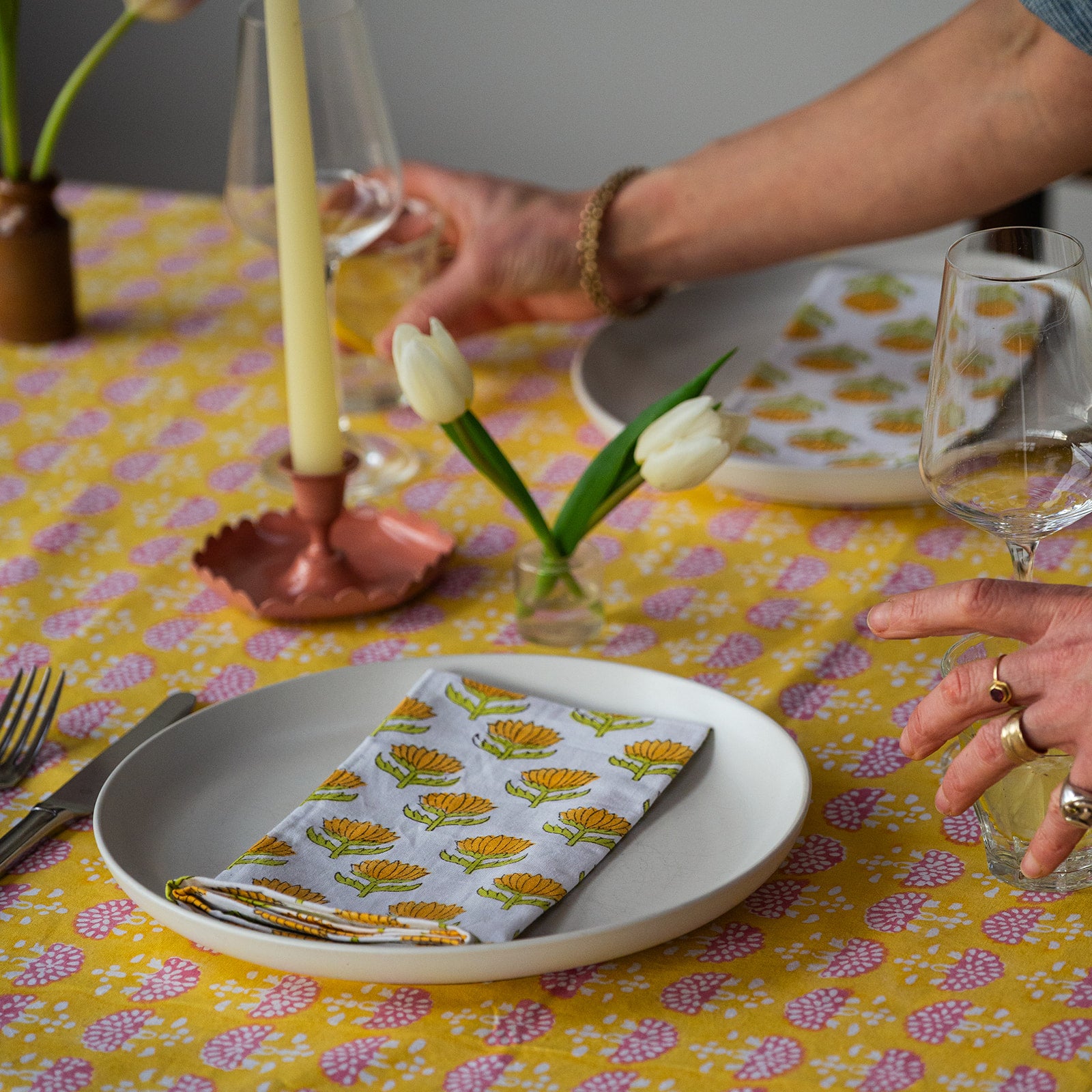 Person setting a table with floral-patterned napkins and candles on a yellow tablecloth.