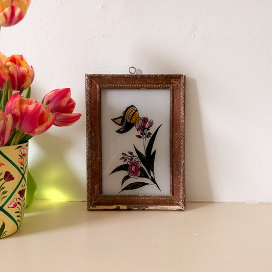 Decorative framed artwork with butterfly and flowers next to a vase of tulips on a white surface.