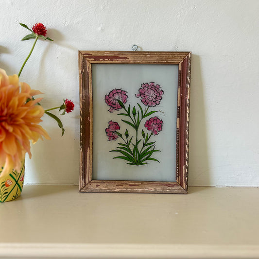 Framed artwork of pink flowers on a white wall with a vase of flowers to the left. constance-and-denny-indian-reverse-glass-painting-boxed-packaging-hand-painted-artisan-art-carnations
