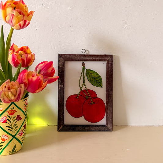 Framed artwork of cherries next to a vase with tulips on a white wall.