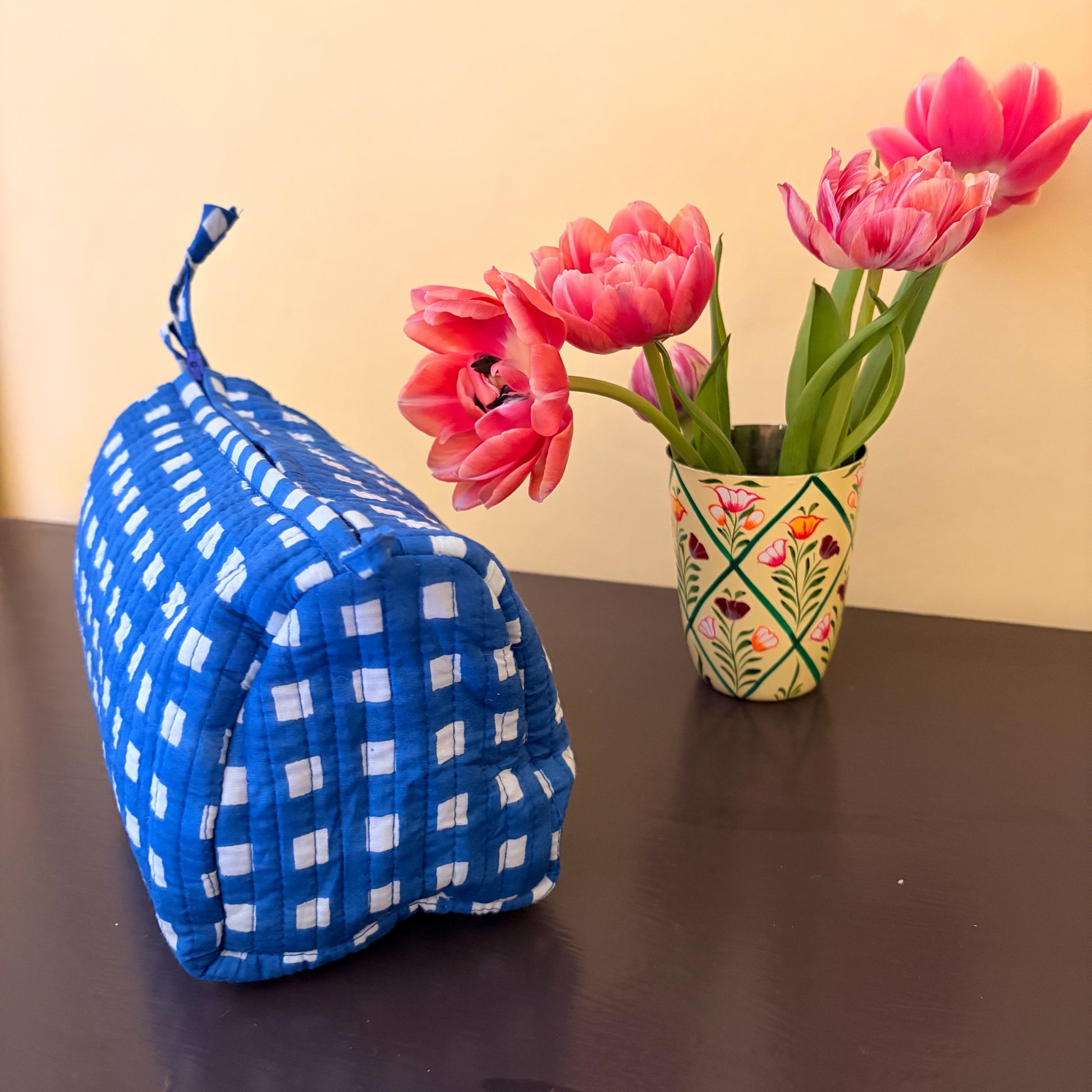 Blue checkered cosmetic bag next to a vase with pink tulips on a dark surface. curved 