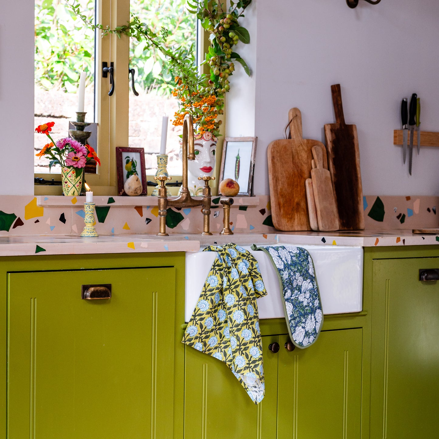 Kitchen with green cabinets, floral tea towels, oven gloves and wooden cutting boards.