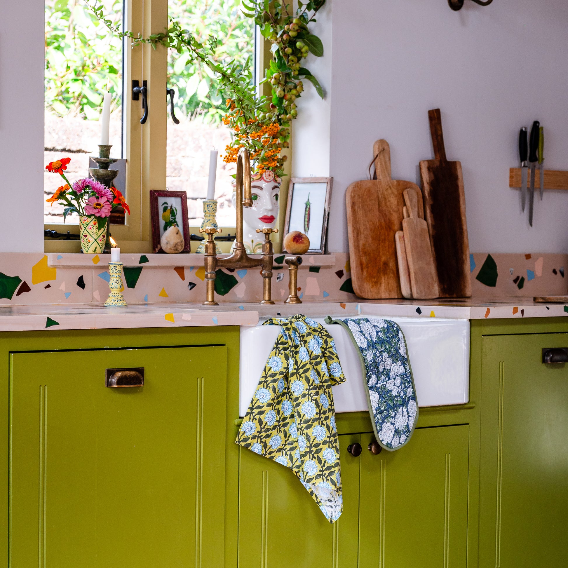 Kitchen with green cabinets, floral tea towels, oven gloves and wooden cutting boards.