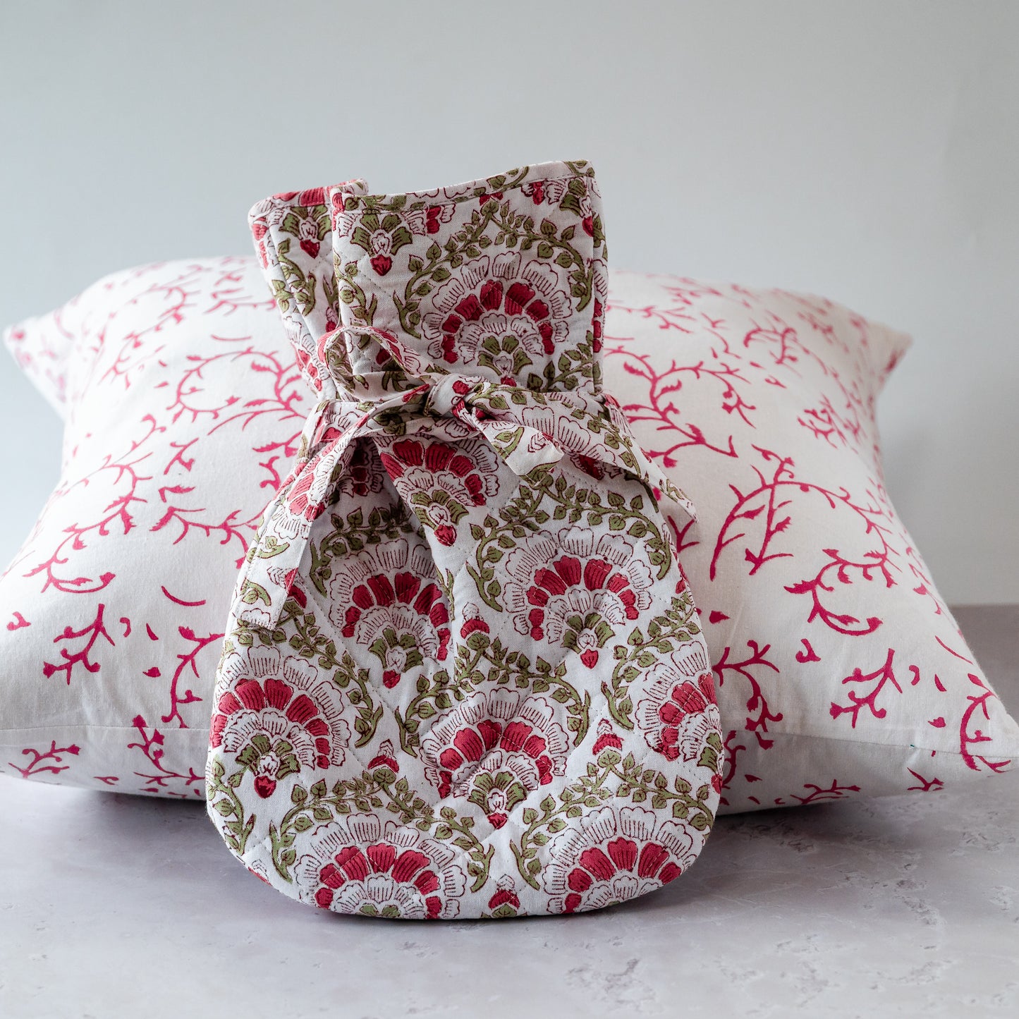 Fabric hot water bottle with floral pattern in front of floral-patterned pillows on a light background