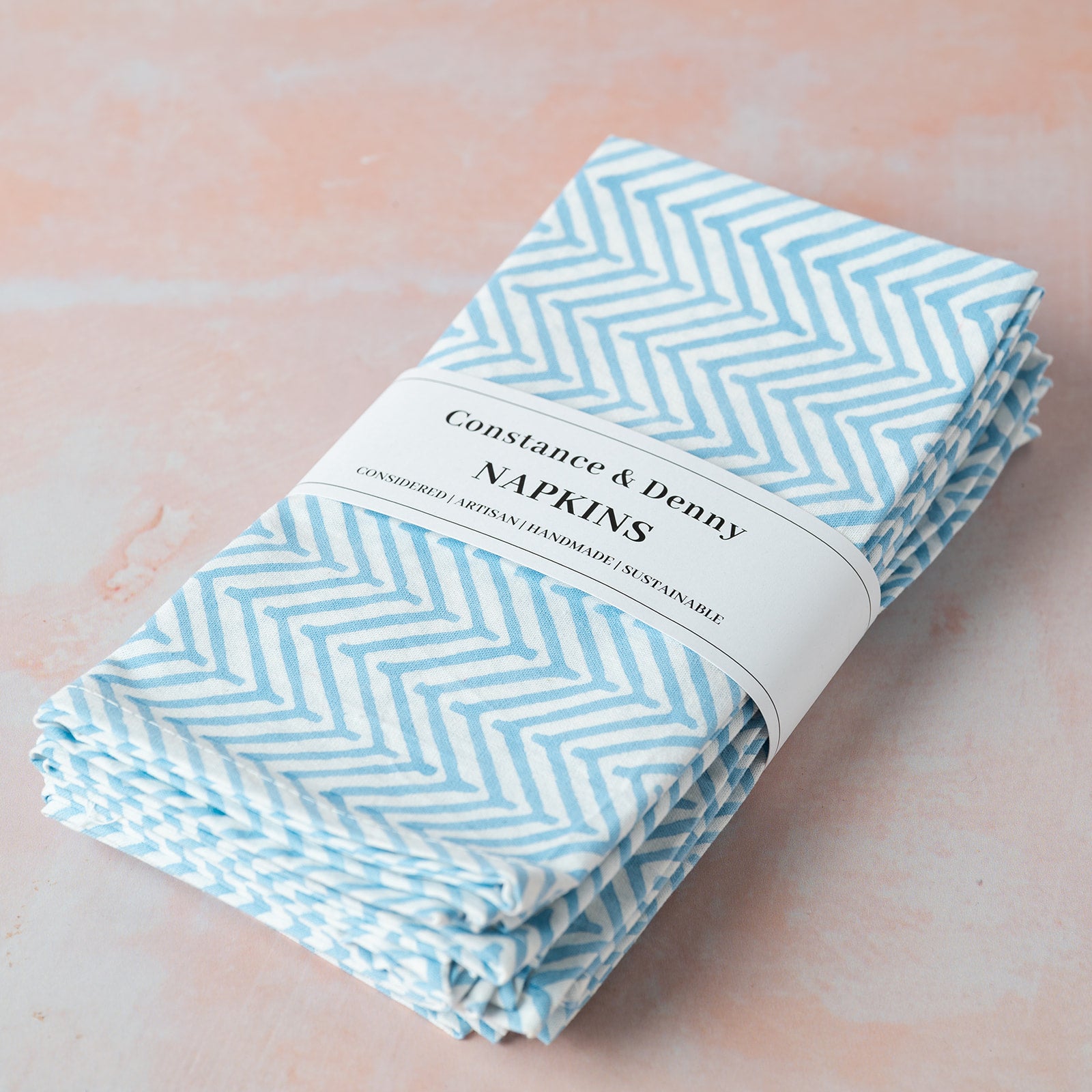 Pack of Constance & Denny napkins with blue chevron pattern on a light pink background