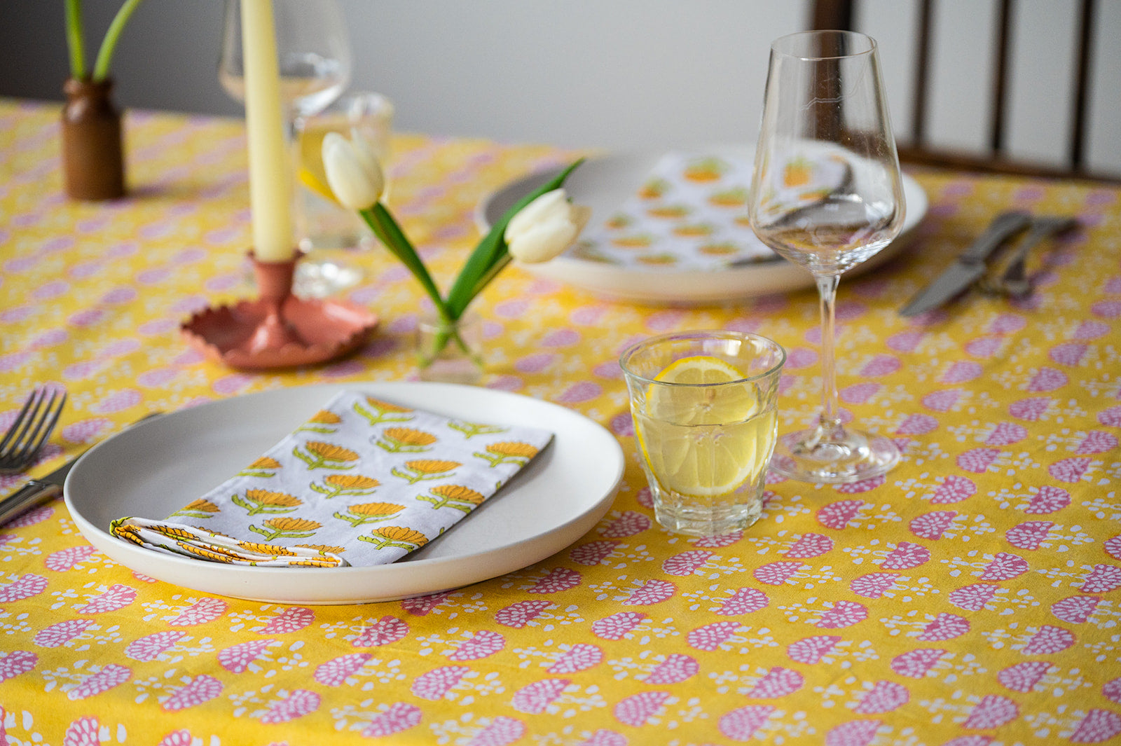 Dining table setting with plates, glasses, and cutlery on a yellow floral tablecloth.