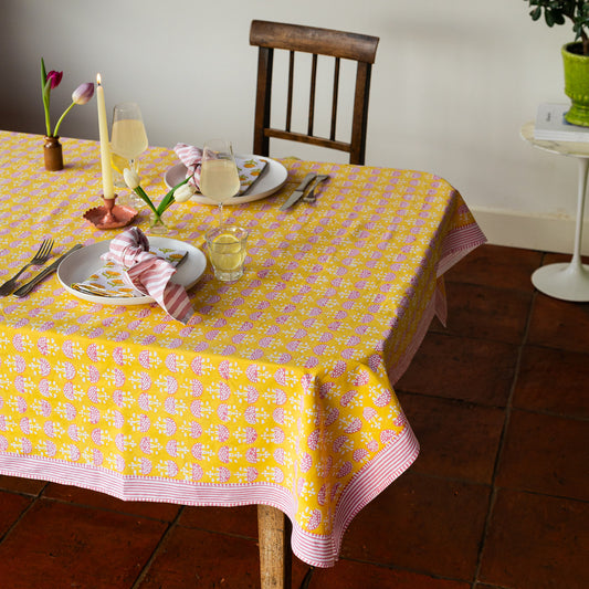 Dining table set with a yellow and pink patterned tablecloth, plates, cutlery, and glasses.