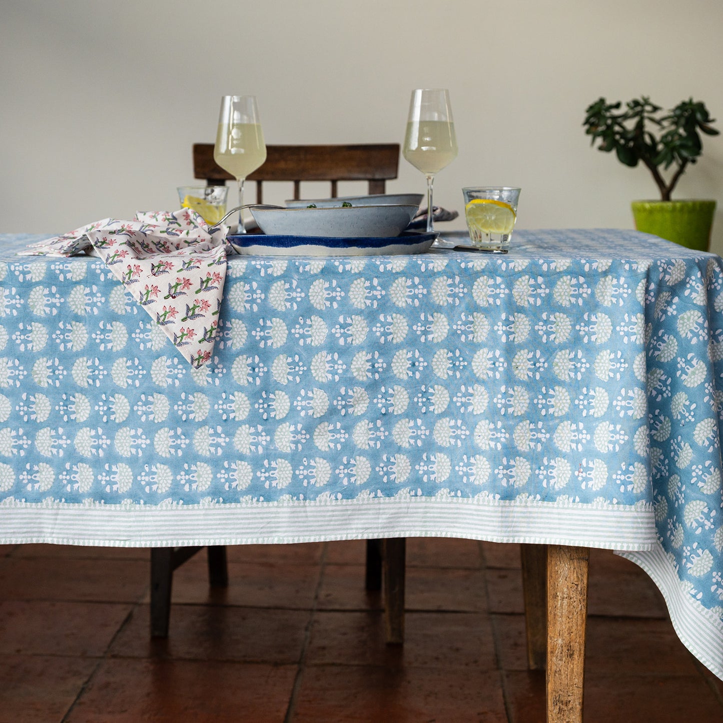 Tablecloth with floral pattern on a dining table with glasses and a plant in the background.