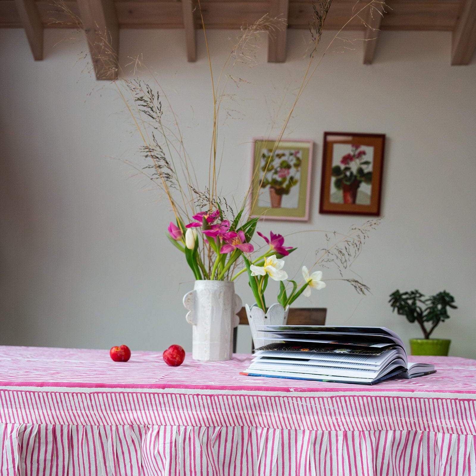 Table with a pink and white checkered tablecloth, flowers in a vase, apples, and magazines.